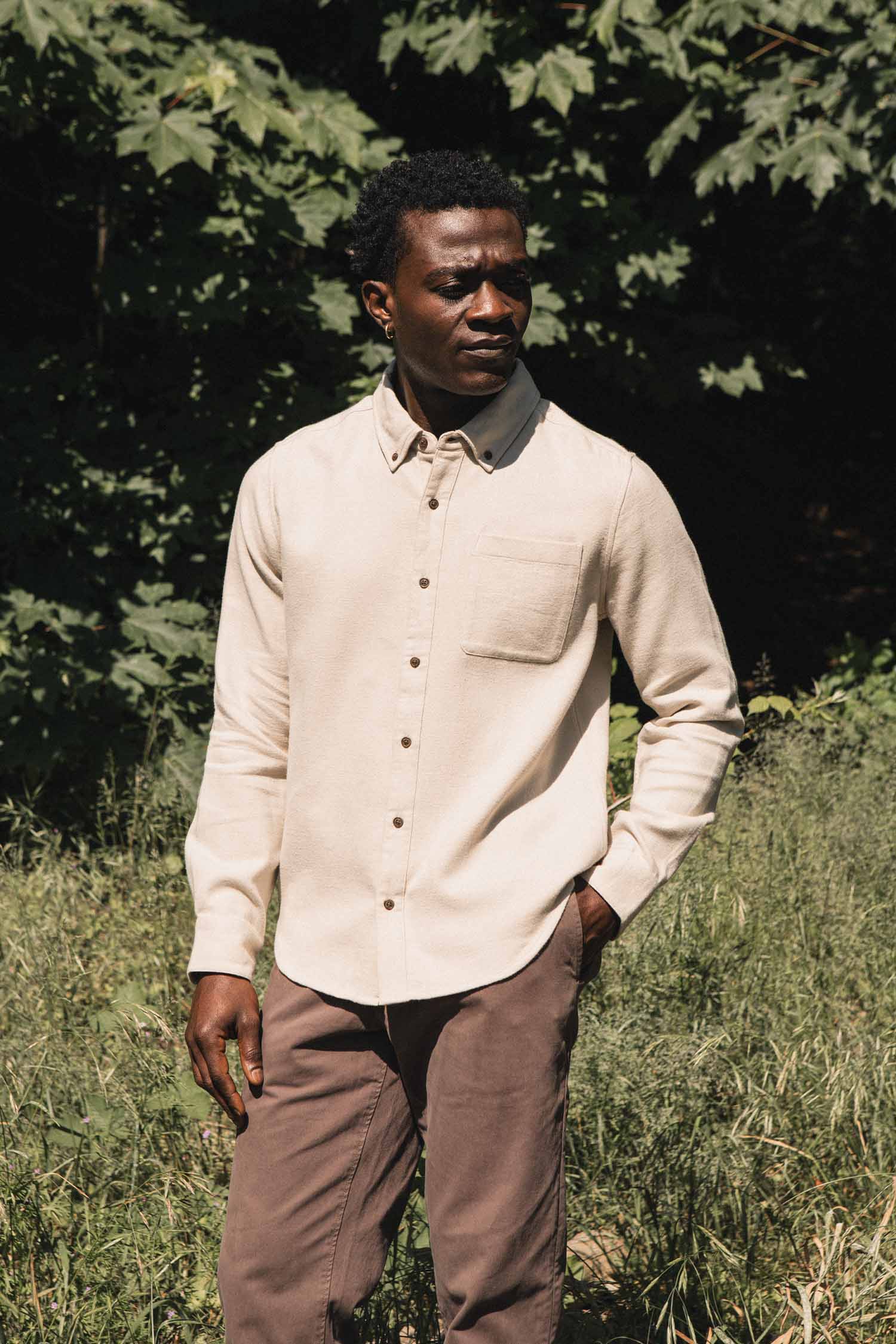 A man stands outdoors in front of green foliage, wearing the Bridge & Burn Sutton Slim Shirt in Fawn Flannel and brown pants, with one hand in his pocket, looking off to the side.