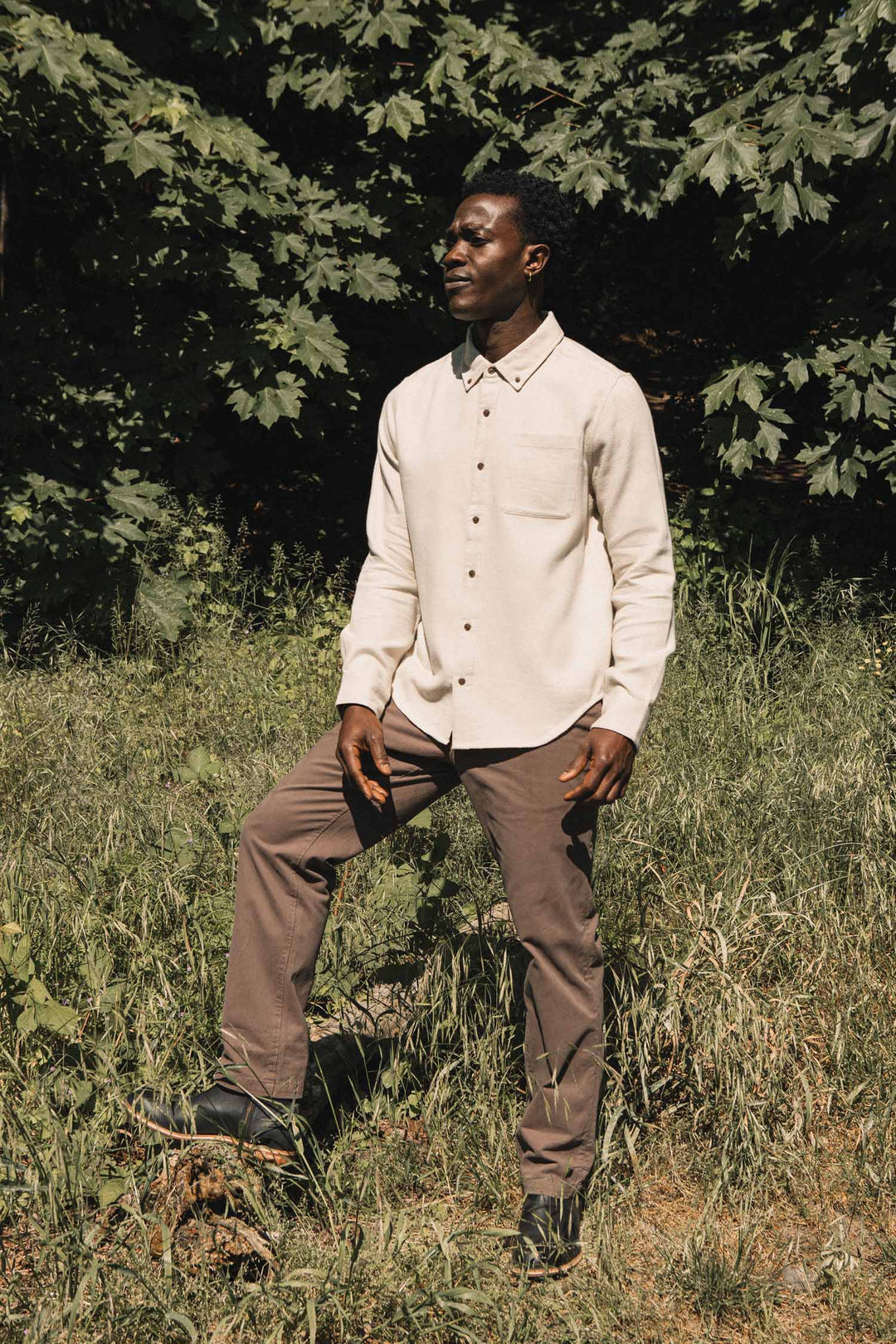 A man stands outdoors on grass with one foot raised, wearing the Bridge & Burn Sutton Slim Shirt in Fawn Flannel and brown pants. Lush green trees and sunlight fill the background.