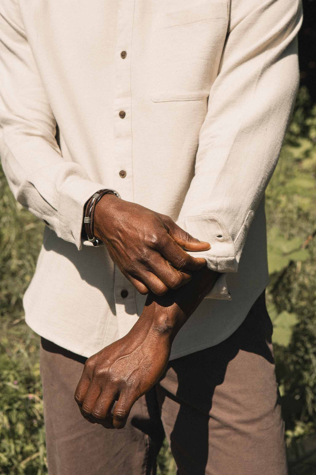 A person wearing the Bridge & Burn Sutton Slim Shirt in Fawn Flannel stands outdoors, adjusting their shirt cuff. The minimalist image highlights their hands and torso, with the cream-toned organic cotton shirt and brown pants set against a softly blurred green background.