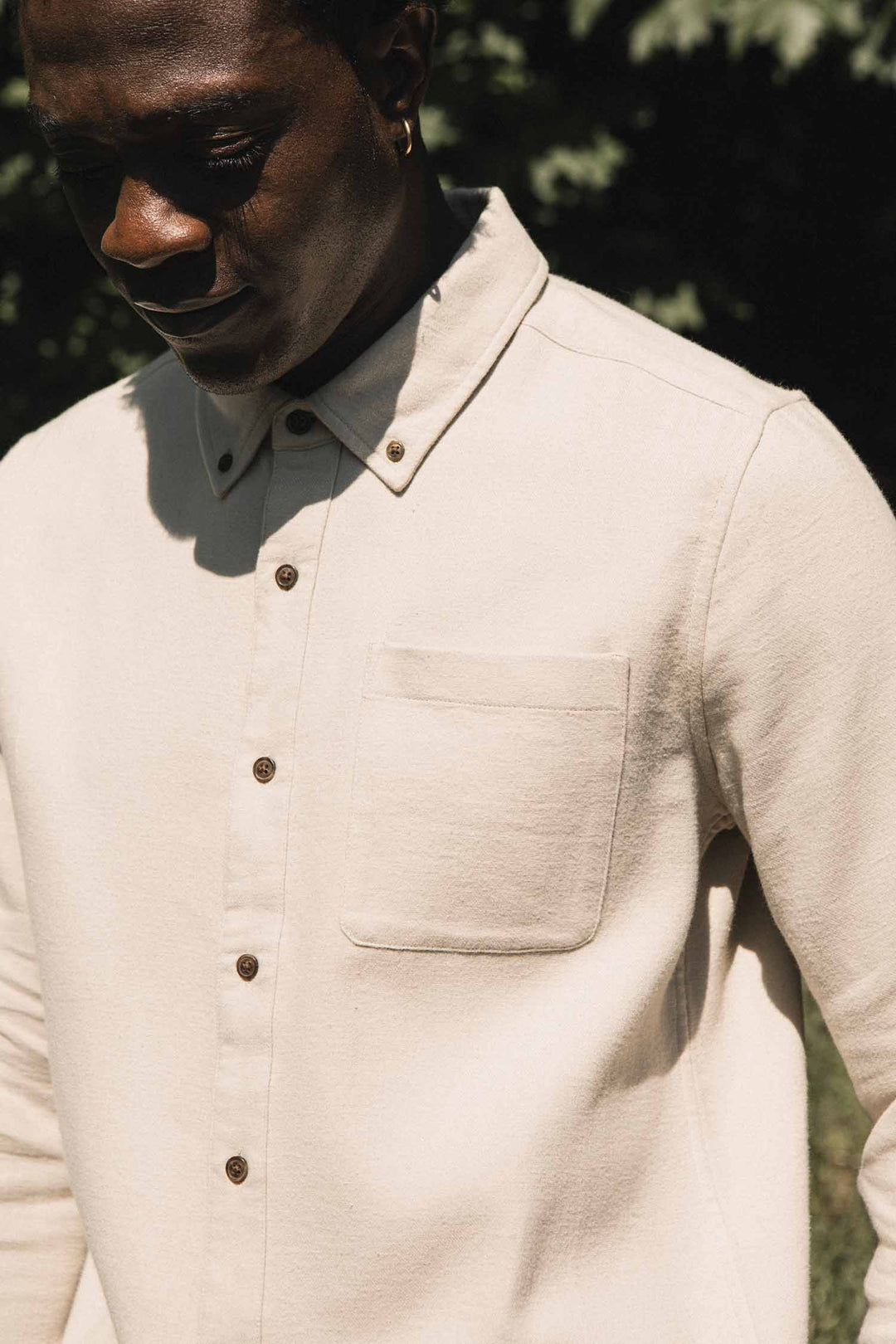 A person stands outdoors in sunlight, their face partially in shadow, embodying a minimalist design while wearing the Sutton Slim Shirt in Fawn Flannel by Bridge & Burn—an organic cotton shirt featuring a chest pocket.