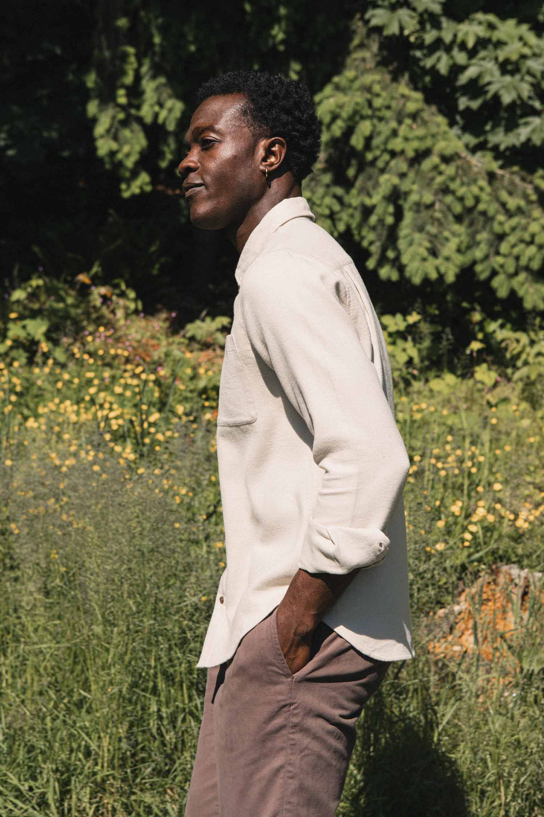 Dressed in the Bridge & Burn Sutton Slim Shirt in Fawn Flannel and brown pants, a person stands outdoors amid green grass, yellow flowers, and trees, reflecting a minimalist aesthetic.