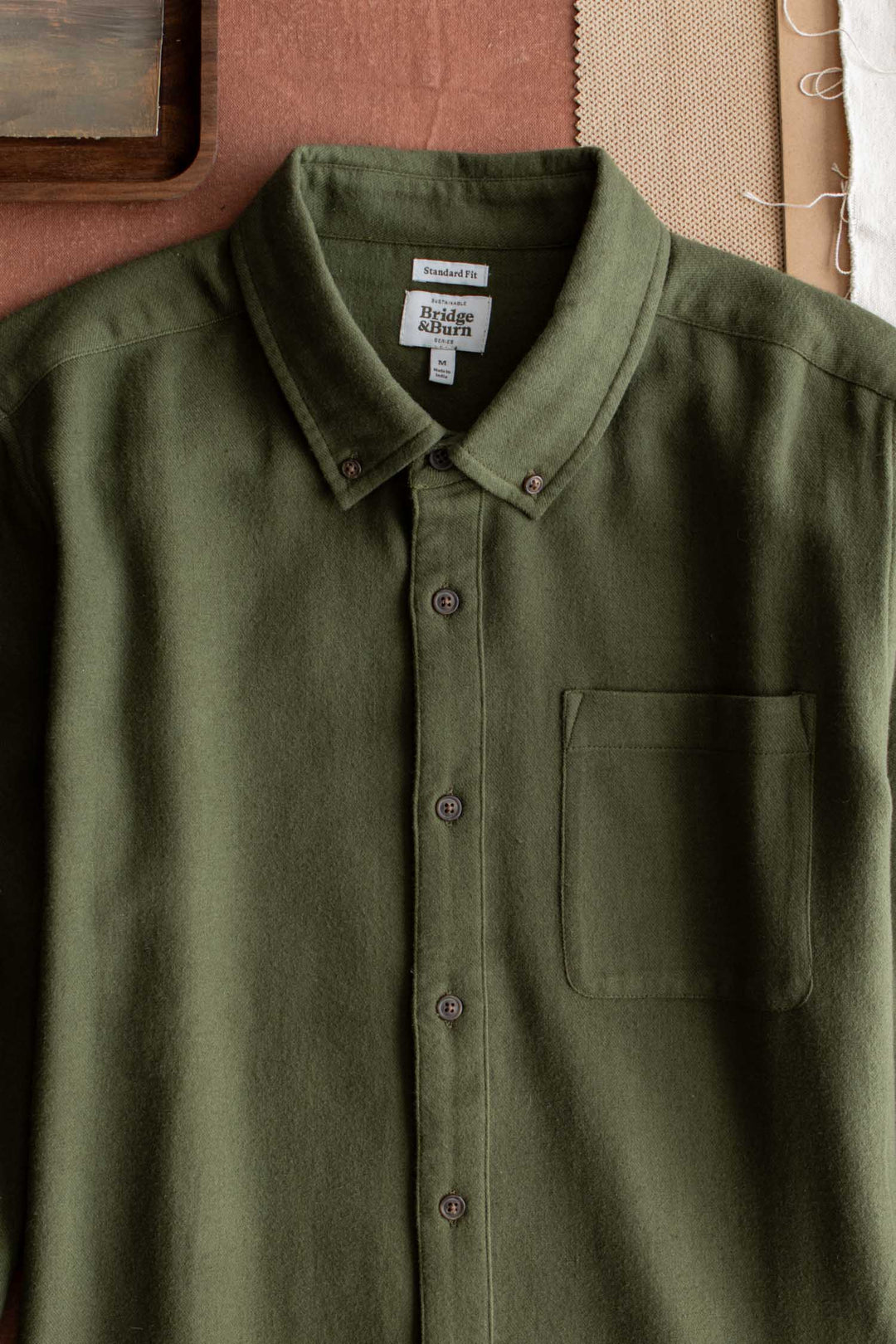 The Bridge & Burn Sutton Slim Shirt in Pine Flannel is laid flat on a brown surface, showcasing its olive green hue and single chest pocket. Crafted from soft, slightly textured organic cotton for comfort and sustainability, this button-up features a buttoned-down collar that emphasizes its minimalist design. Fabric swatches are arranged in the background.