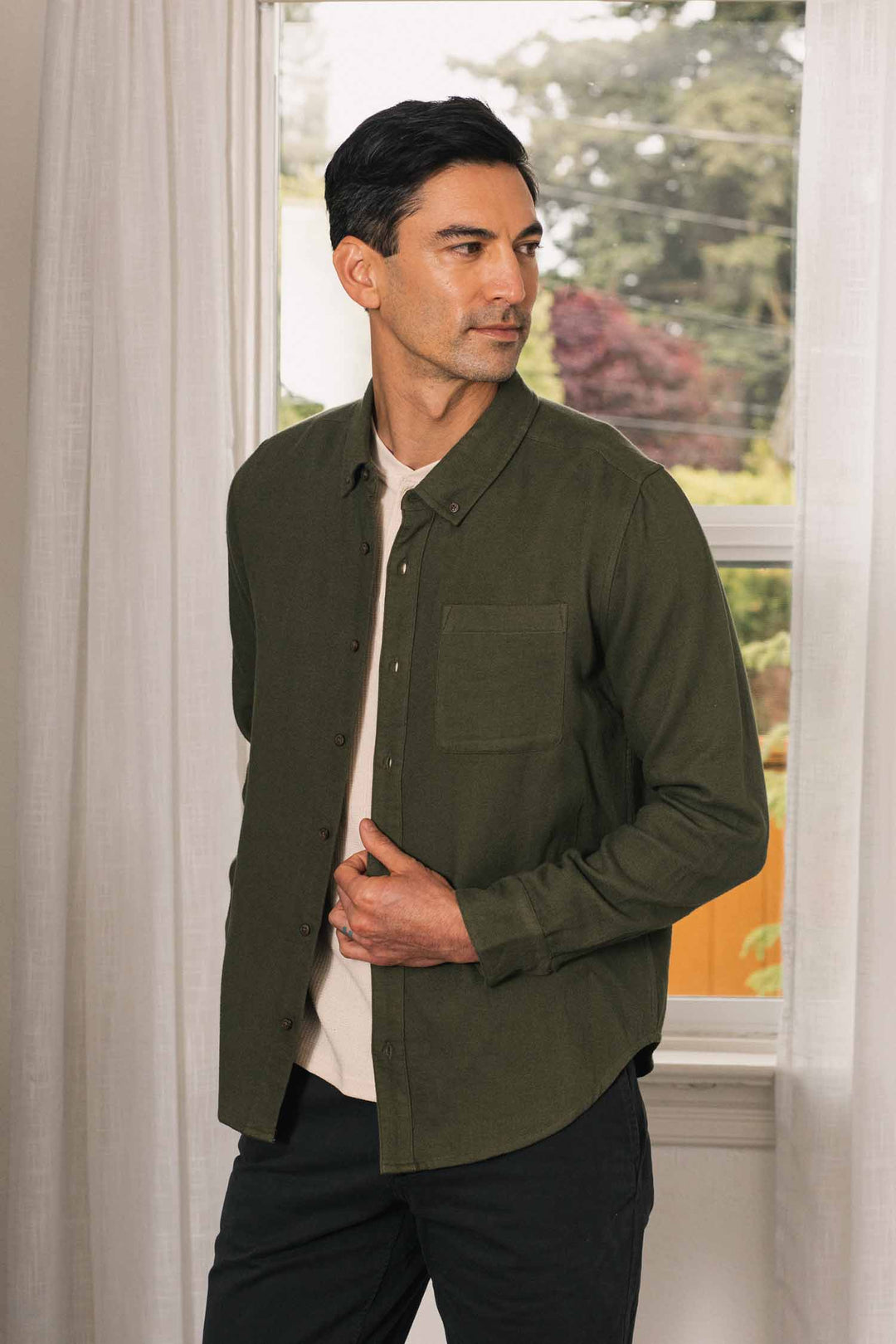 A man stands indoors by a window, wearing the Bridge & Burn Sutton Slim Shirt in Pine Flannel over a light-colored t-shirt and black pants. He looks to his left, adjusting his shirt with one hand. Trees are visible outside the window, complementing the space’s minimalist design.