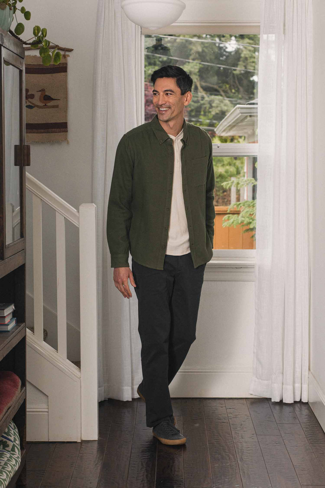 A man stands smiling in a well-lit hallway near a window, wearing the Bridge & Burn Sutton Slim Shirt in pine flannel over a light shirt, paired with black pants and dark shoes. The cozy minimalist space features stairs, bookshelves, and indoor plants.