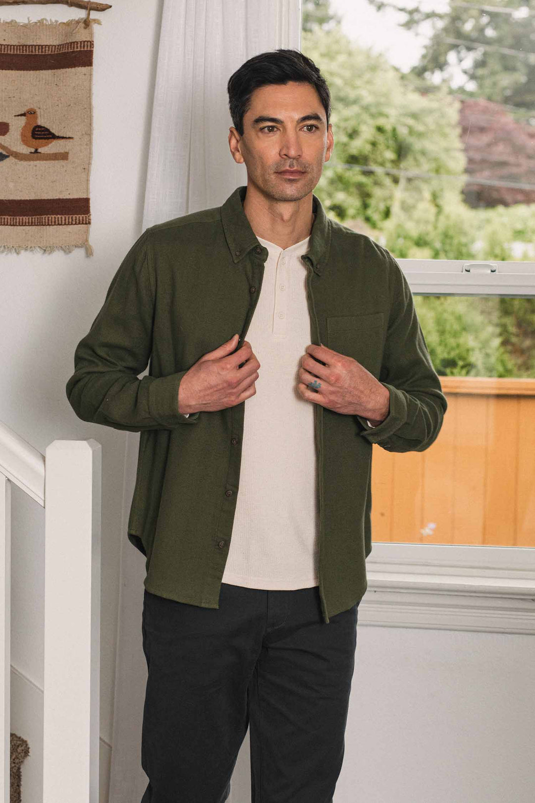 A man stands indoors by a window, wearing the Bridge & Burn Sutton Slim Shirt in Pine Flannel over a white henley and dark pants, adjusting his shirt with both hands. A beige wall hanging and a staircase are nearby.