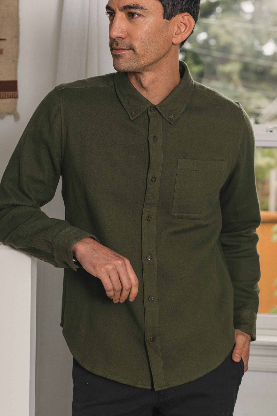 A man wearing the Bridge & Burn Sutton Slim Shirt in Pine Flannel, featuring an olive green slim fit and a chest pocket, stands indoors near a window, looking to the side with one hand resting on a table and the other in his pocket.