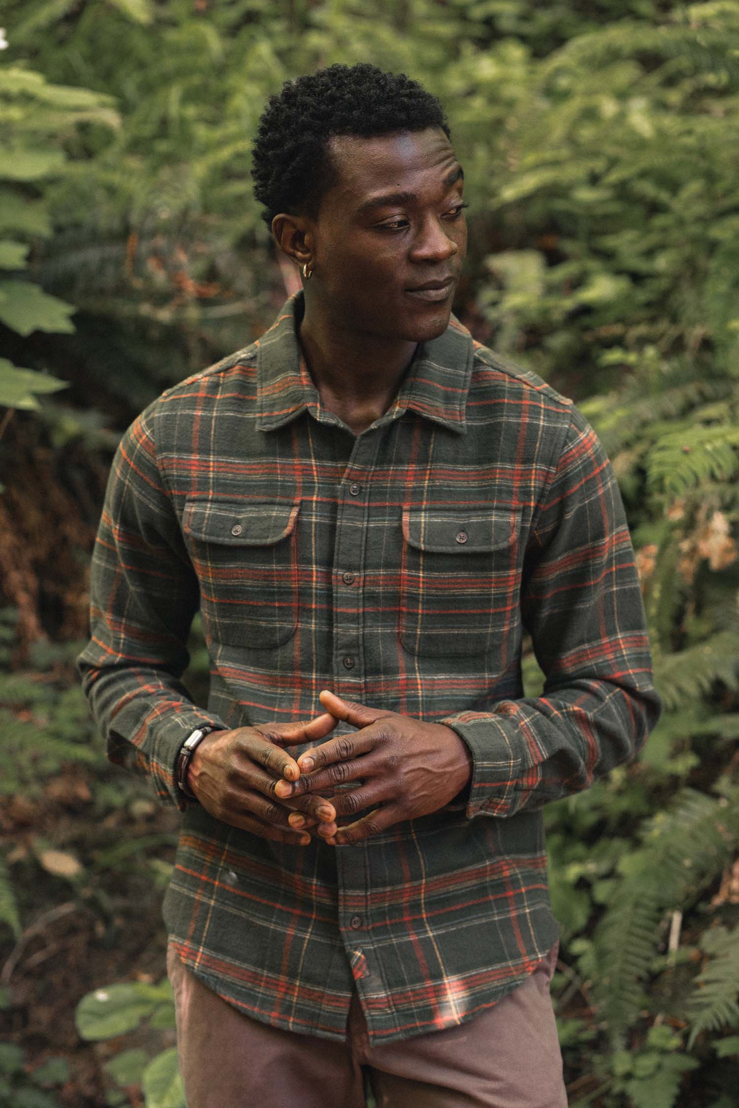 A man wearing a green and orange Bridge & Burn Cole Slim Shirt / Timberline Plaid button-down with two chest pockets, standing in front of greenery.