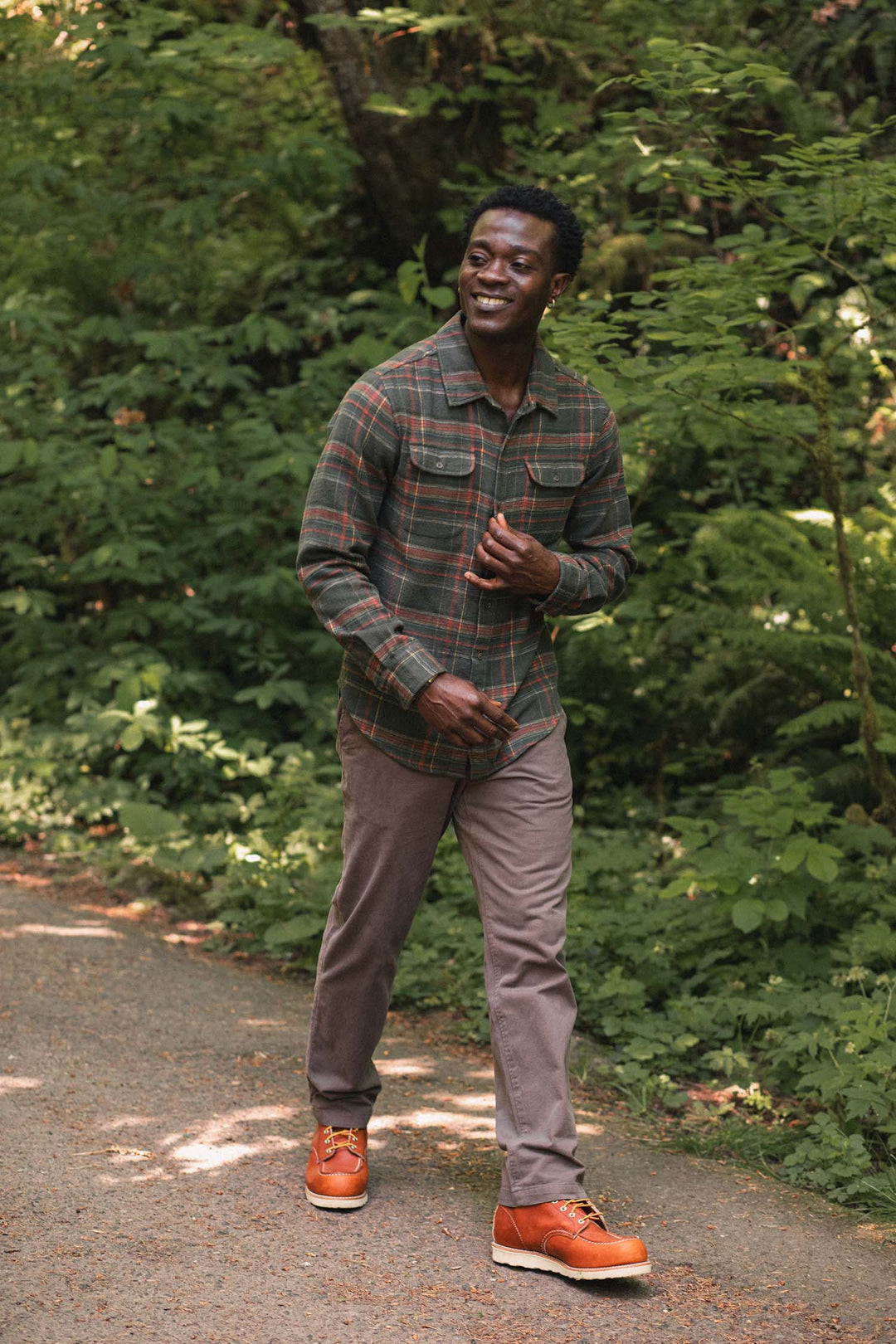 A man wearing a green and red plaid Cole Slim Shirt / Timberline Plaid by Bridge & Burn and brown pants walks along a forest path.