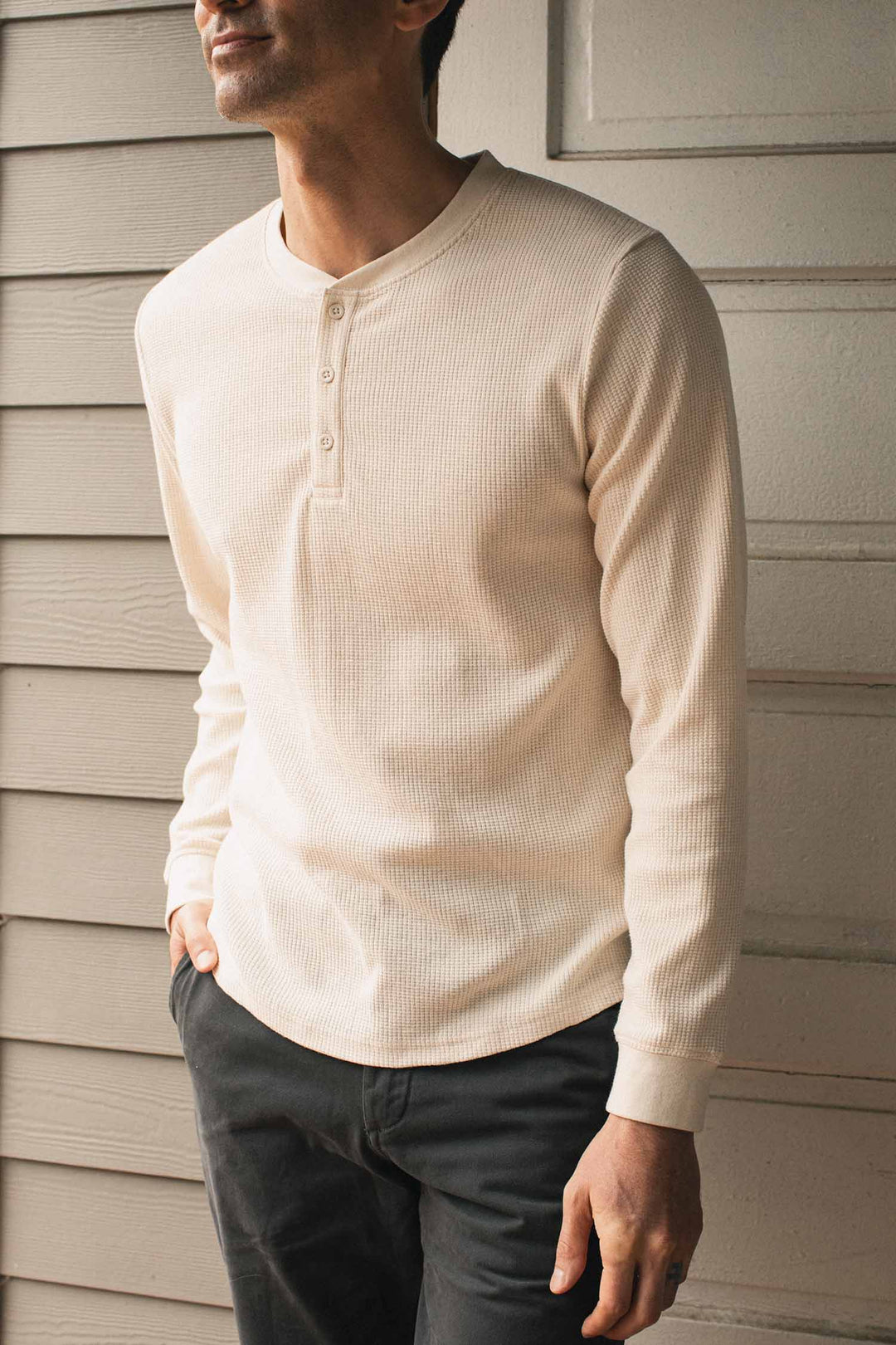 A person wearing the Bridge & Burn Men’s Recycled Waffle Henley in Ivory and dark pants stands in front of a beige, paneled wall, with hands relaxed by their sides.