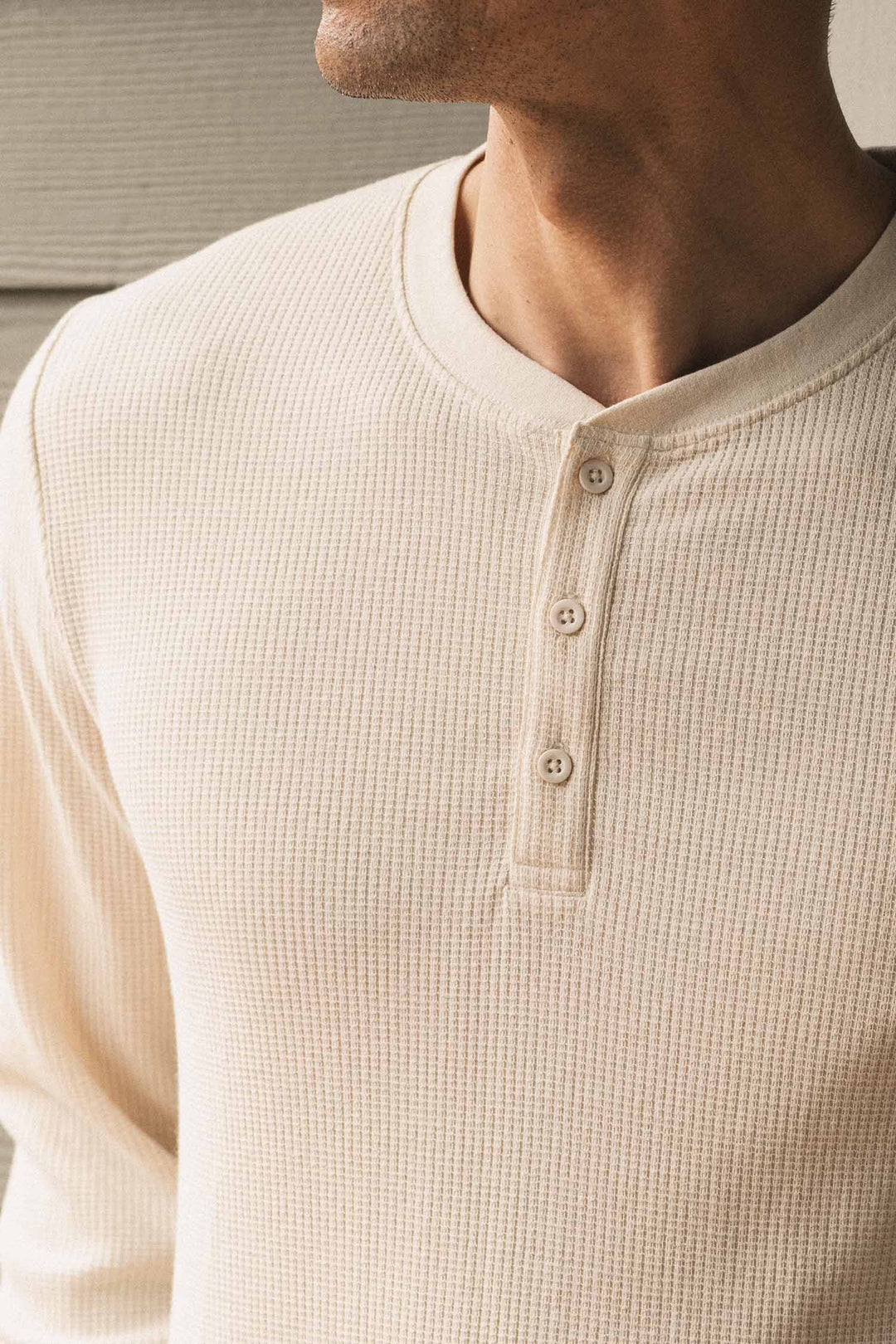 A man is shown wearing the Bridge & Burn Men’s Recycled Waffle Henley in Ivory, a cream-colored, long-sleeve thermal layering shirt with a three-button collar. Only the lower half of his face and upper torso are visible.