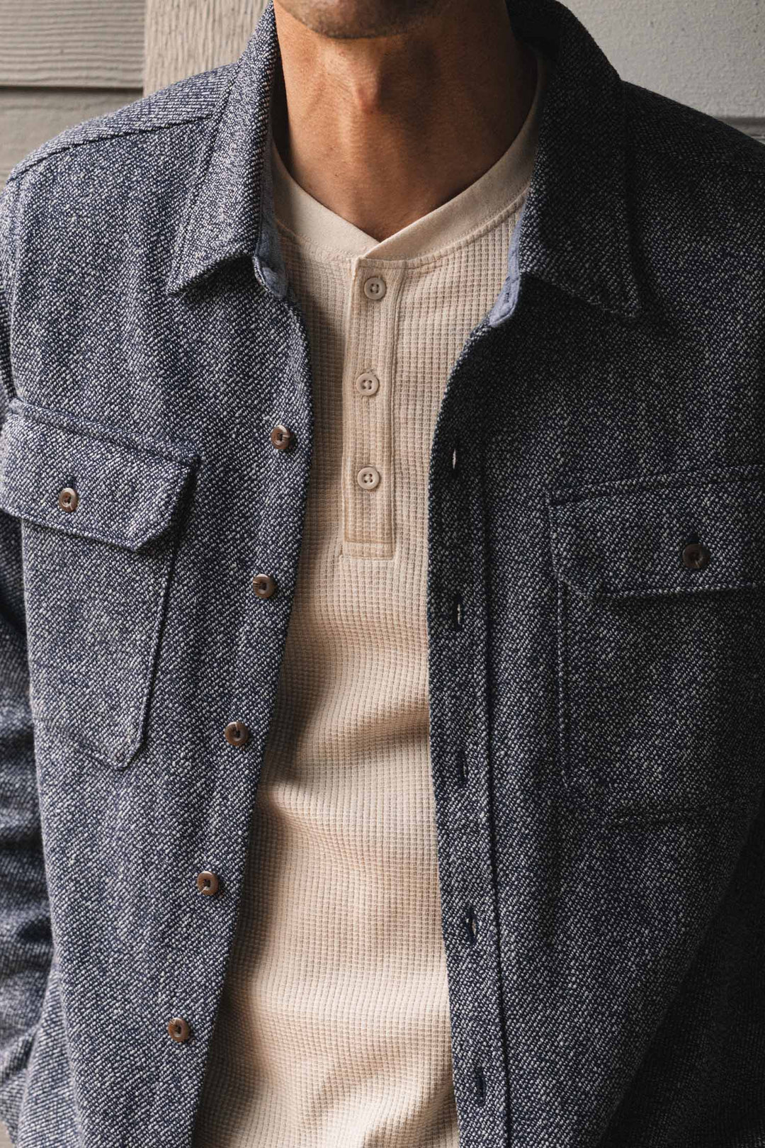 A man wearing the Men’s Recycled Waffle Henley in Ivory by Bridge & Burn, featuring three buttons and a textured knit, layered under a blue button-up jacket; shown from shoulders to waist against a neutral background.
