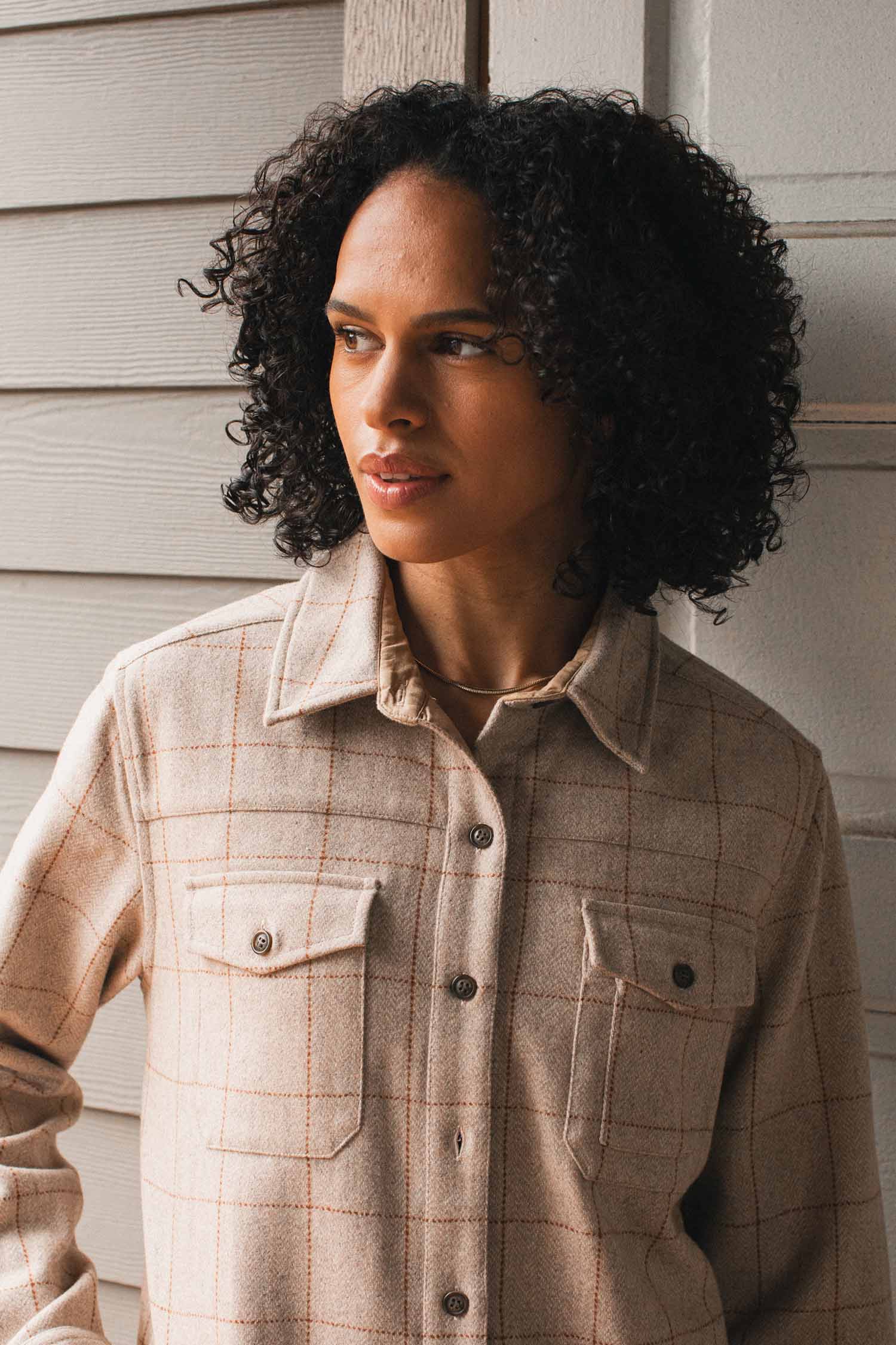 A person with curly dark hair wears the Bridge & Burn Harlow Shirt Jacket in Stonecross, standing before a light wooden wall and gazing thoughtfully to the side.