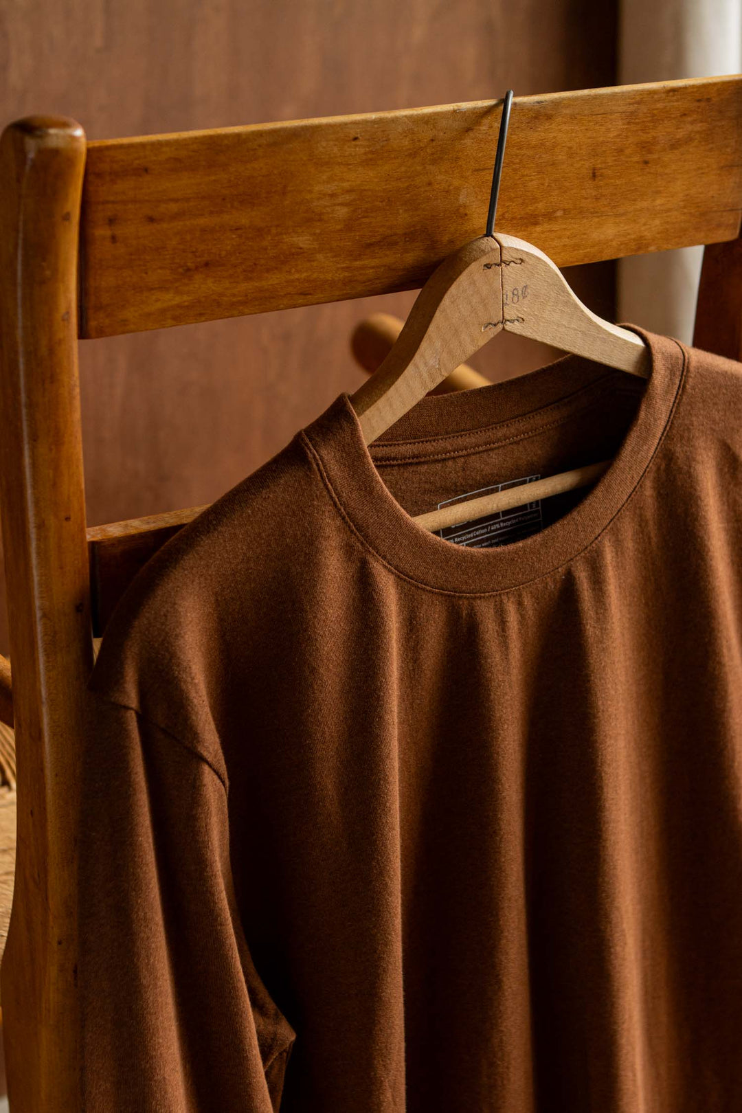 The Audrey Long Sleeve Tee in Bronze by Bridge & Burn hangs on a wooden hanger over the back of a wooden chair, illuminated by warm indoor lighting against a softly blurred brown background. Its relaxed fit enhances the cozy feel of the scene.