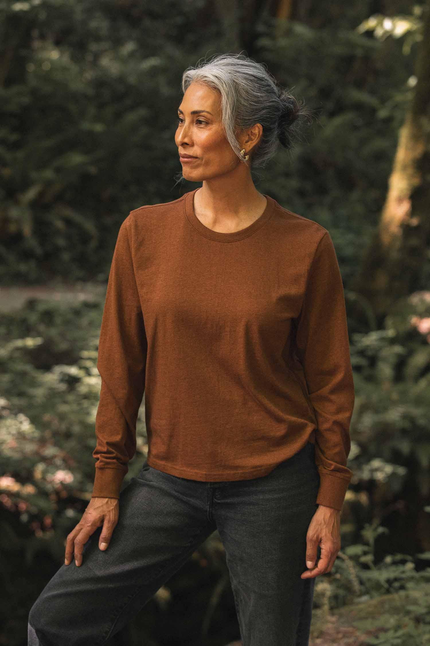A woman with gray hair pulled back wears a Bridge & Burn Audrey Long Sleeve Tee in Bronze, crafted from soft jersey knit, paired with dark jeans as she stands outdoors surrounded by a lush, green forest. The relaxed fit of her shirt complements her calm expression as she looks to the side.