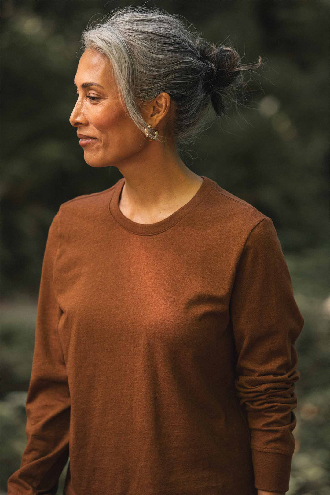 A woman with gray hair in a bun, wearing the Bridge & Burn Audrey Long Sleeve Tee in Bronze—a relaxed fit jersey knit—stands outdoors, looking to the left with a gentle smile.