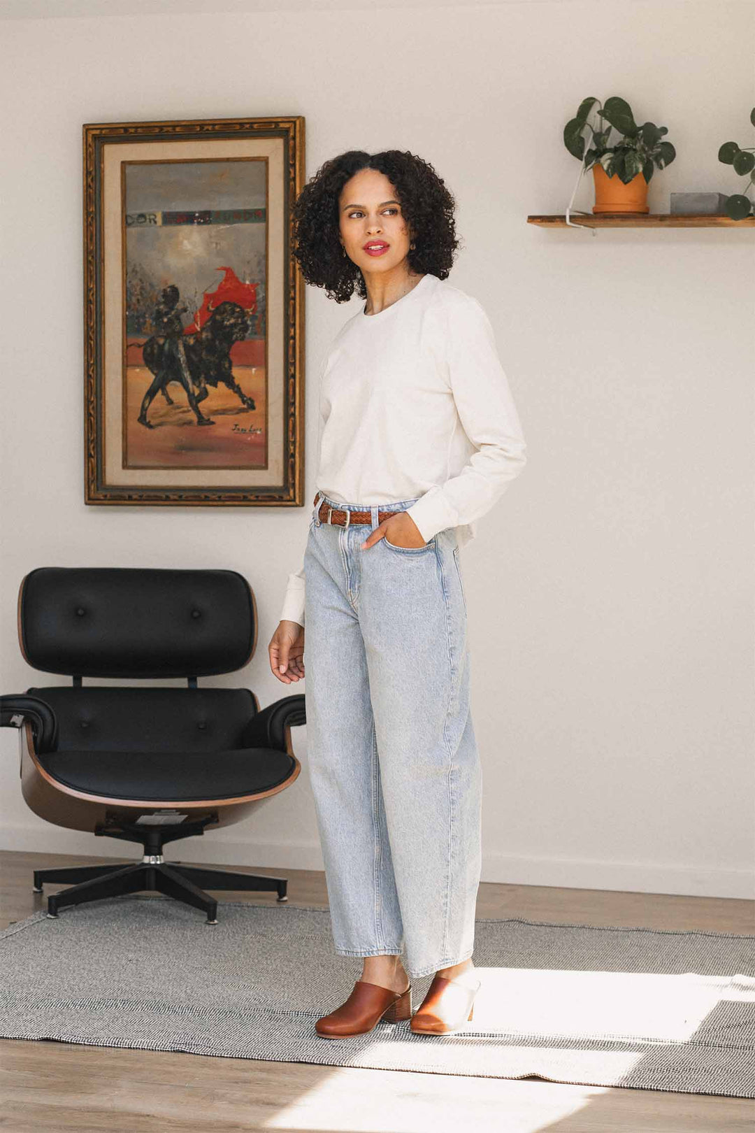 A woman stands indoors on a rug, wearing a relaxed fit Audrey Long Sleeve Tee in Ivory by Bridge & Burn, made from recycled cotton, paired with light blue jeans and brown shoes. Behind her are a black chair, a framed painting, and a white shelf with potted plants.