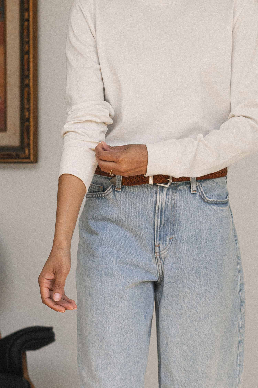 Wearing the Bridge & Burn Audrey Long Sleeve Tee in Ivory, made from recycled cotton, and light blue jeans, a person adjusts their sleeve. Their head is not visible. A brown braided belt and a portion of a framed picture are also in view, emphasizing the relaxed fit of their outfit.