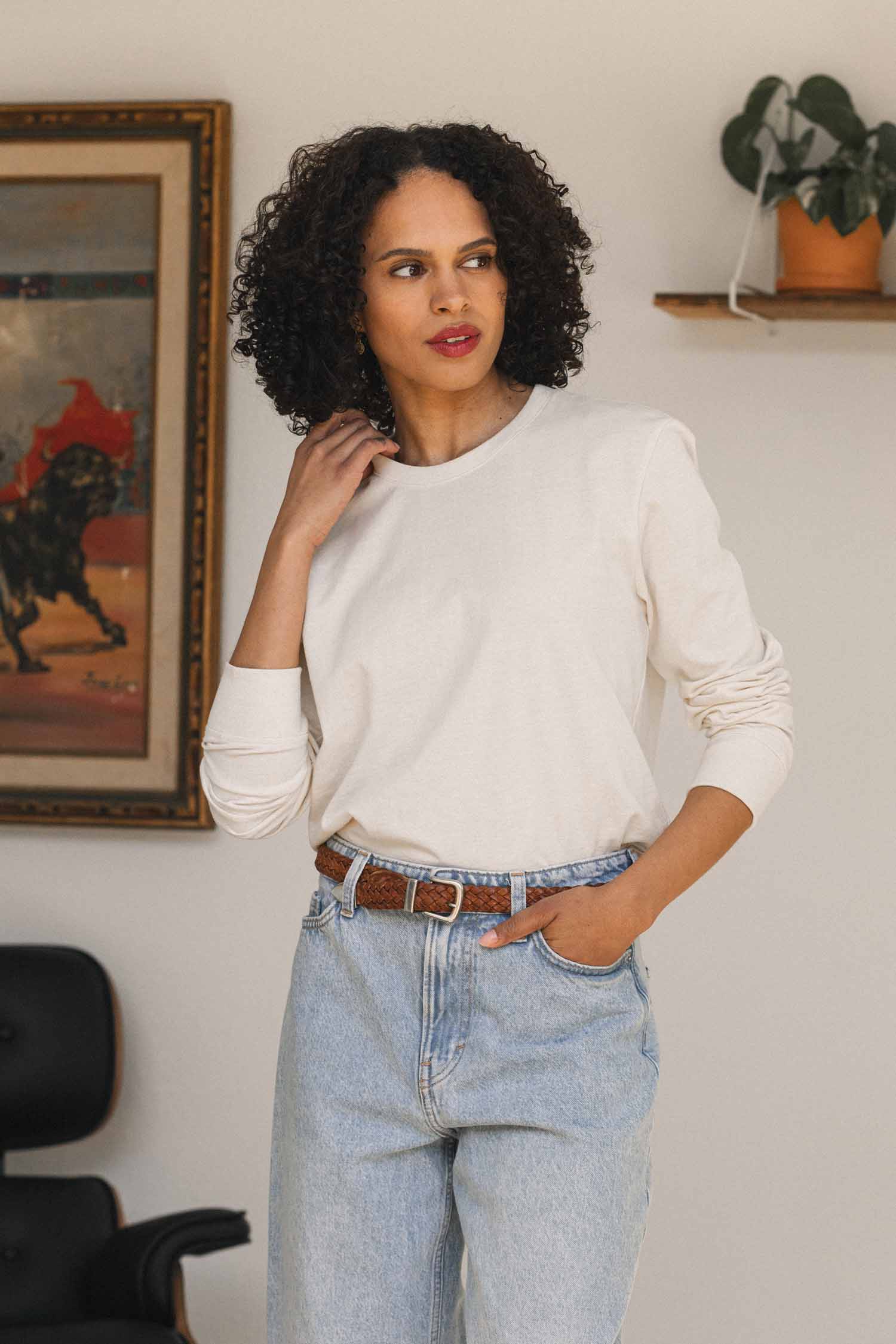 A woman with curly hair stands indoors, her hand gently touching her neck. She wears the Bridge & Burn Audrey Long Sleeve Tee in Ivory, crafted from recycled cotton, paired with light blue jeans. The relaxed fit of the shirt enhances her casual look. In the background are a painting, a shelf with a plant, and a black chair.