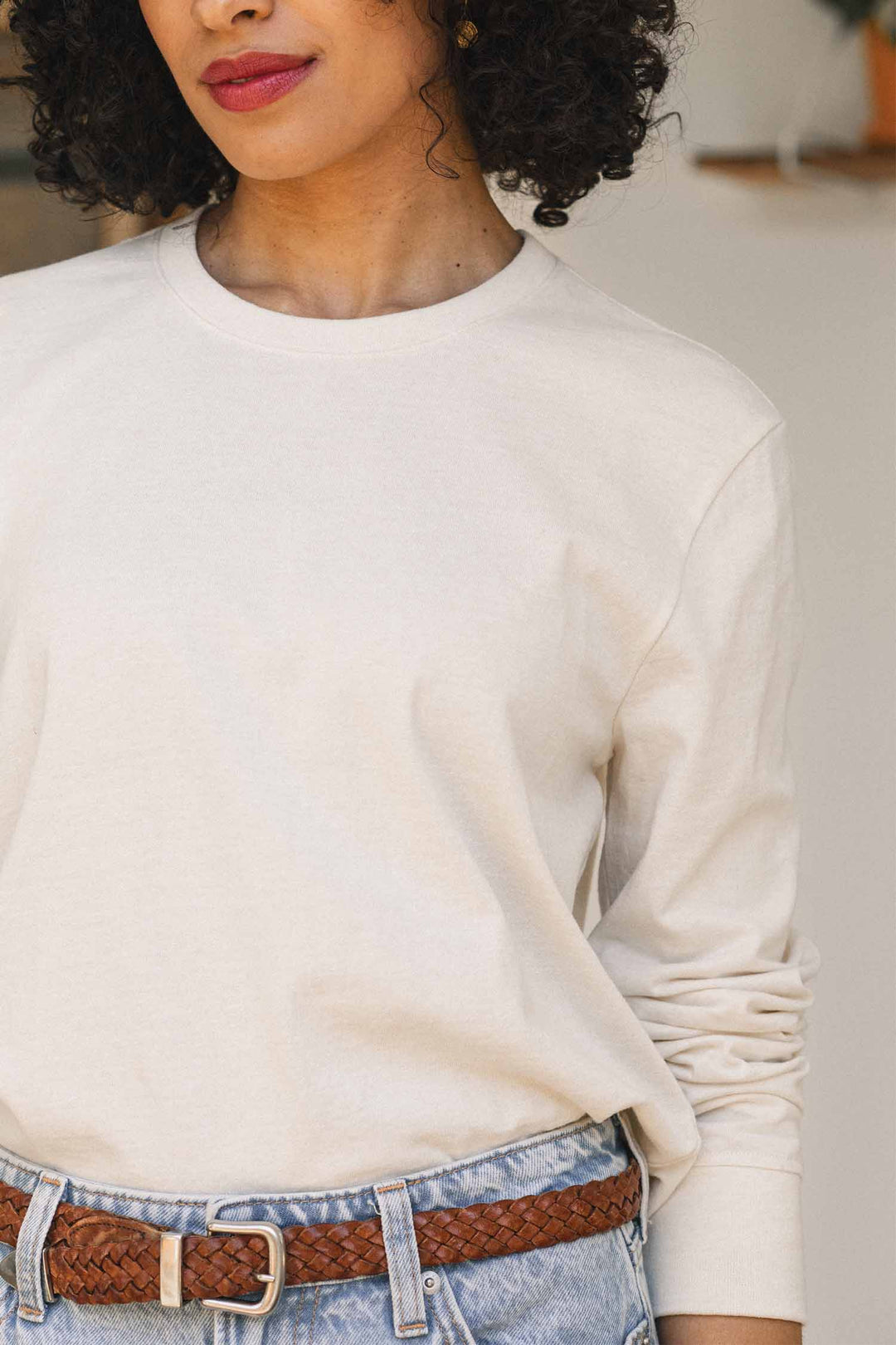 A woman wearing the Bridge & Burn Audrey Long Sleeve Tee in Ivory, paired with light blue recycled cotton jeans and a brown braided belt, stands with her hands in her pockets. Only her upper body is visible.
