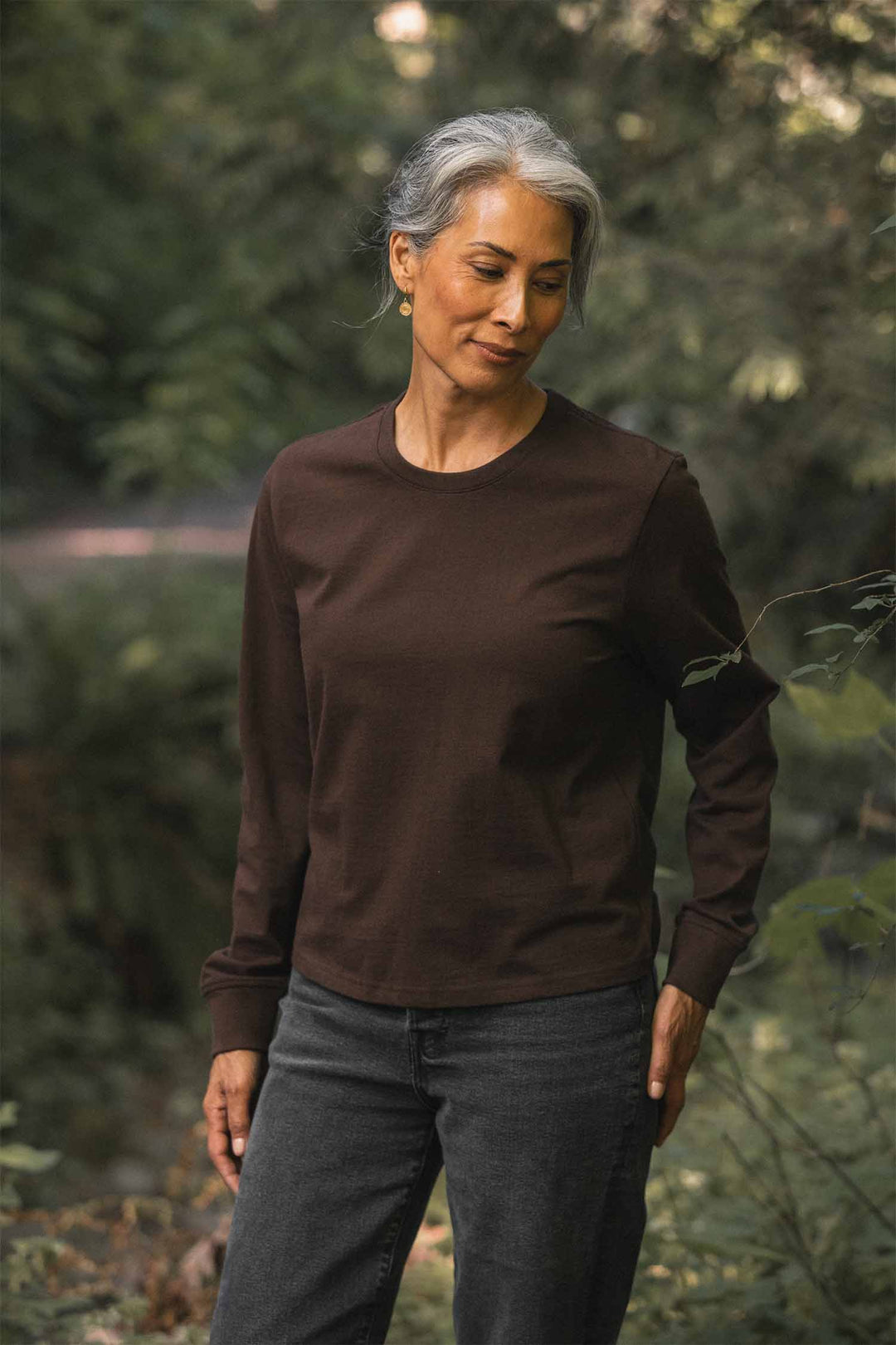A person with gray hair, wearing a relaxed fit Bridge & Burn Audrey Long Sleeve Tee in Truffle made from recycled cotton and dark jeans, stands outdoors in a lush green forest, looking down and smiling softly.