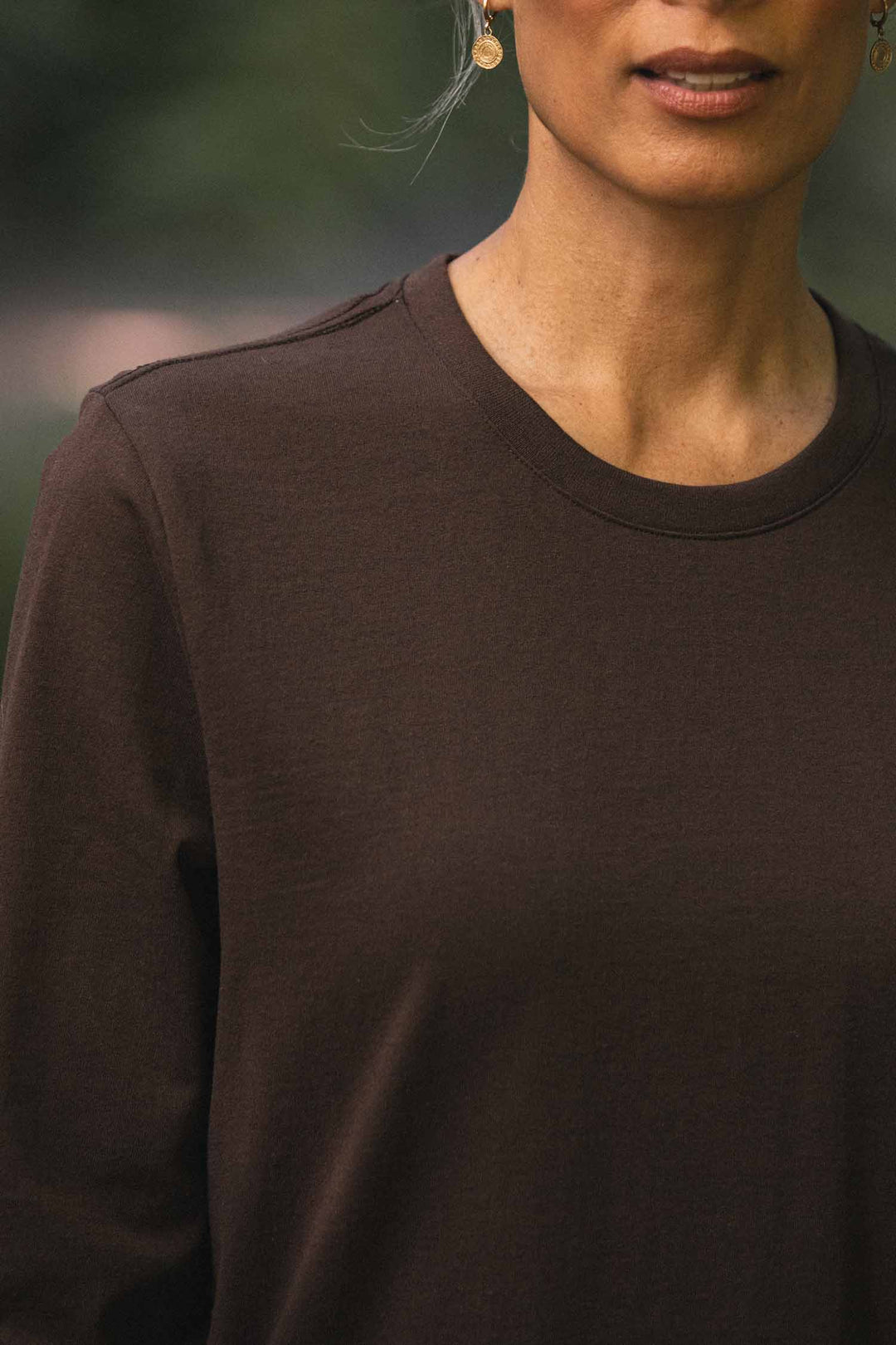 A person is wearing a relaxed fit Bridge & Burn Audrey Long Sleeve Tee in Truffle, made from recycled cotton, along with gold earrings. They are pictured from the shoulders up standing outdoors against a blurred green background.