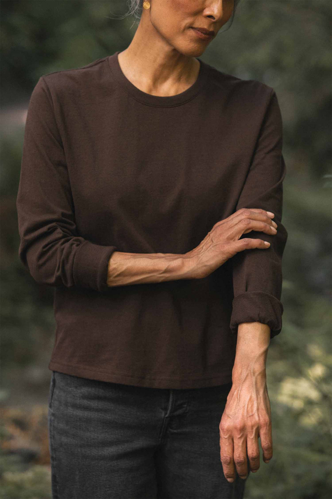 A person wearing the Bridge & Burn Audrey Long Sleeve Tee in Truffle and black jeans stands outdoors, rolling up one sleeve. Their face is partially out of frame, set against a backdrop of blurred greenery. Made from recycled cotton, the relaxed fit tee adds to the effortless style of the scene.