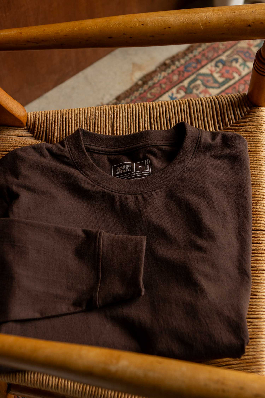 A folded Bridge & Burn Audrey Long Sleeve Tee in Truffle, made from recycled cotton and designed with a relaxed fit, rests on a woven wooden chair, with a glimpse of a patterned rug and wooden floor in the background.