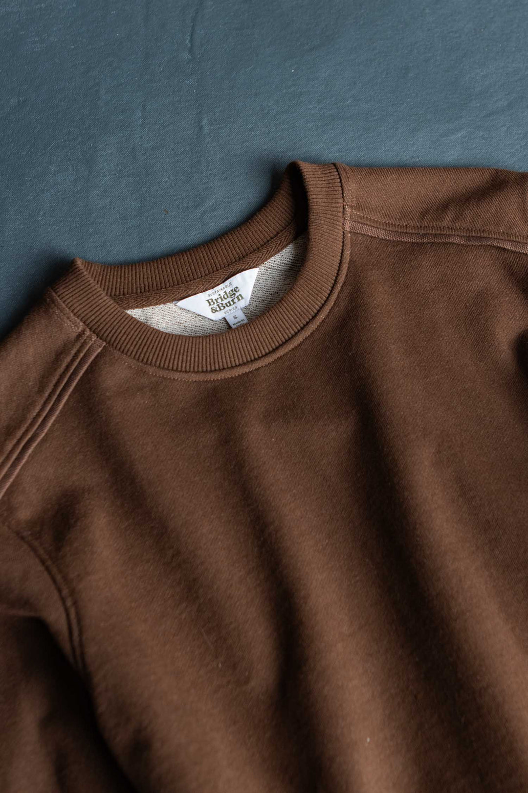 A Bailey Crew Sweatshirt in the cumin colorway by Bridge & Burn is laid flat on a dark surface, displaying its collar, shoulder seams, and part of the sleeve. The neck tag reads Bridge Classic USA.
