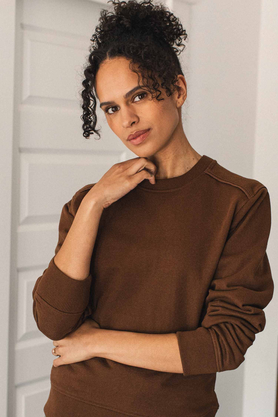 A woman with curly hair stands indoors in front of a white paneled door, looking at the camera with one hand touching her chin and her other arm crossed, wearing the Bridge & Burn Bailey Crew Sweatshirt in Cumin.