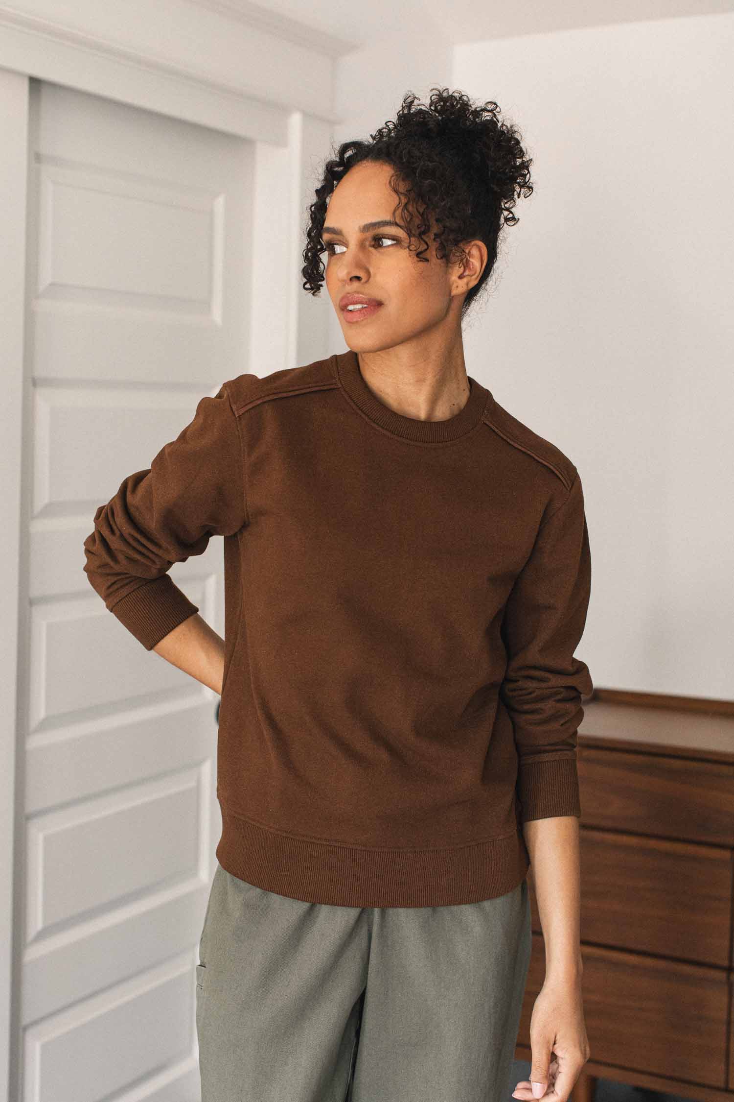 A person with curly hair is wearing the Bridge & Burn Bailey Crew Sweatshirt in Cumin along with gray and brown pants, standing indoors and looking to the side with a neutral expression. White doors and wooden furniture can be seen in the background.
