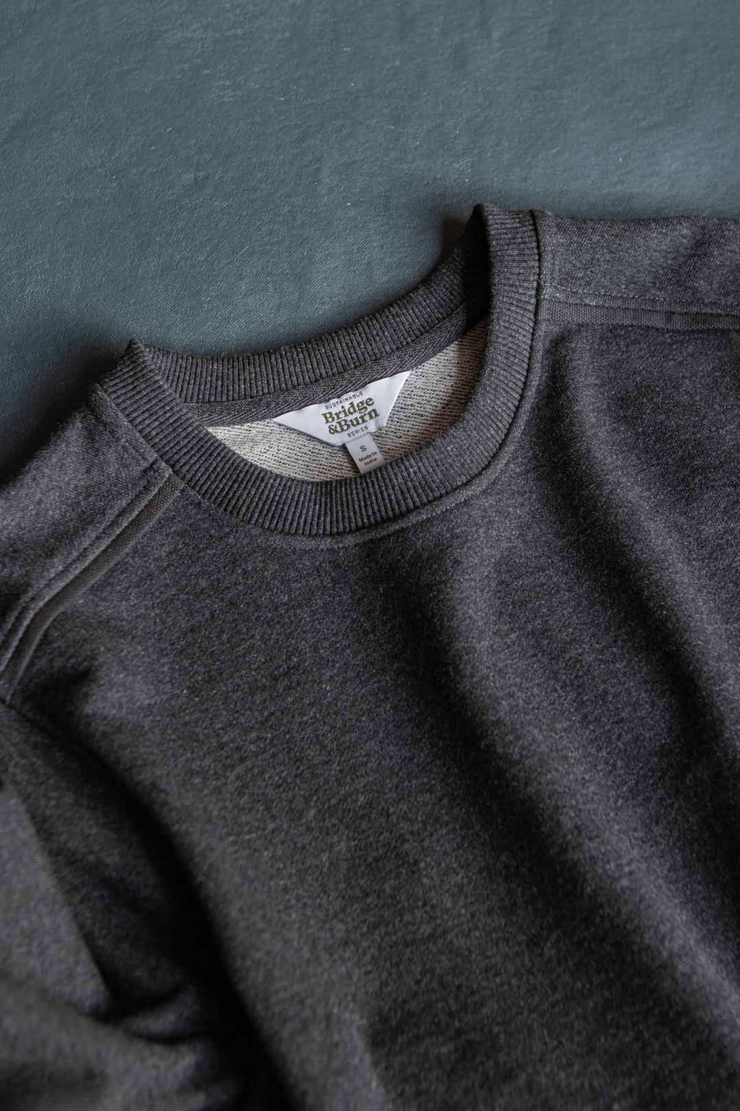 A close-up of the Bridge & Burn Bailey Crew Sweatshirt in Graphite laying flat on a gray surface, highlighting its collar, shoulder seams, and visible neck tag.