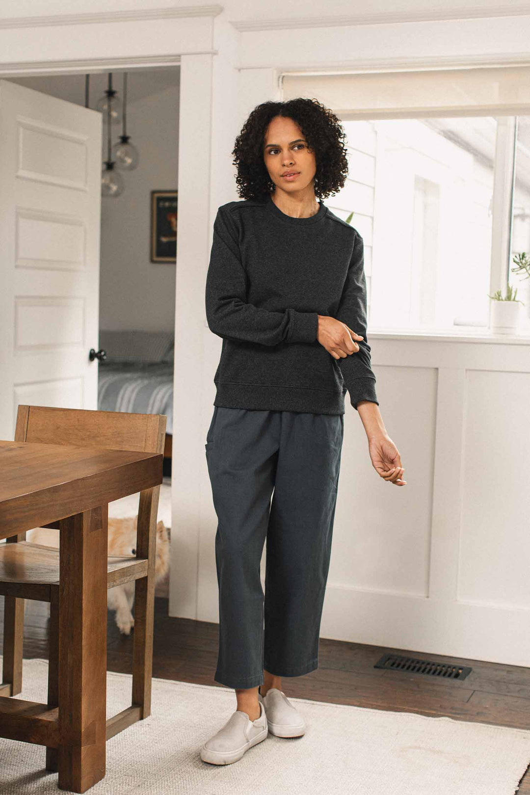 A woman with curly hair, wearing the Bridge & Burn Bailey Crew Sweatshirt in Graphite, blue-gray pants, and white shoes, stands in a bright room next to a wooden table while adjusting her sleeve. A light-colored dog is partially visible in the background.
