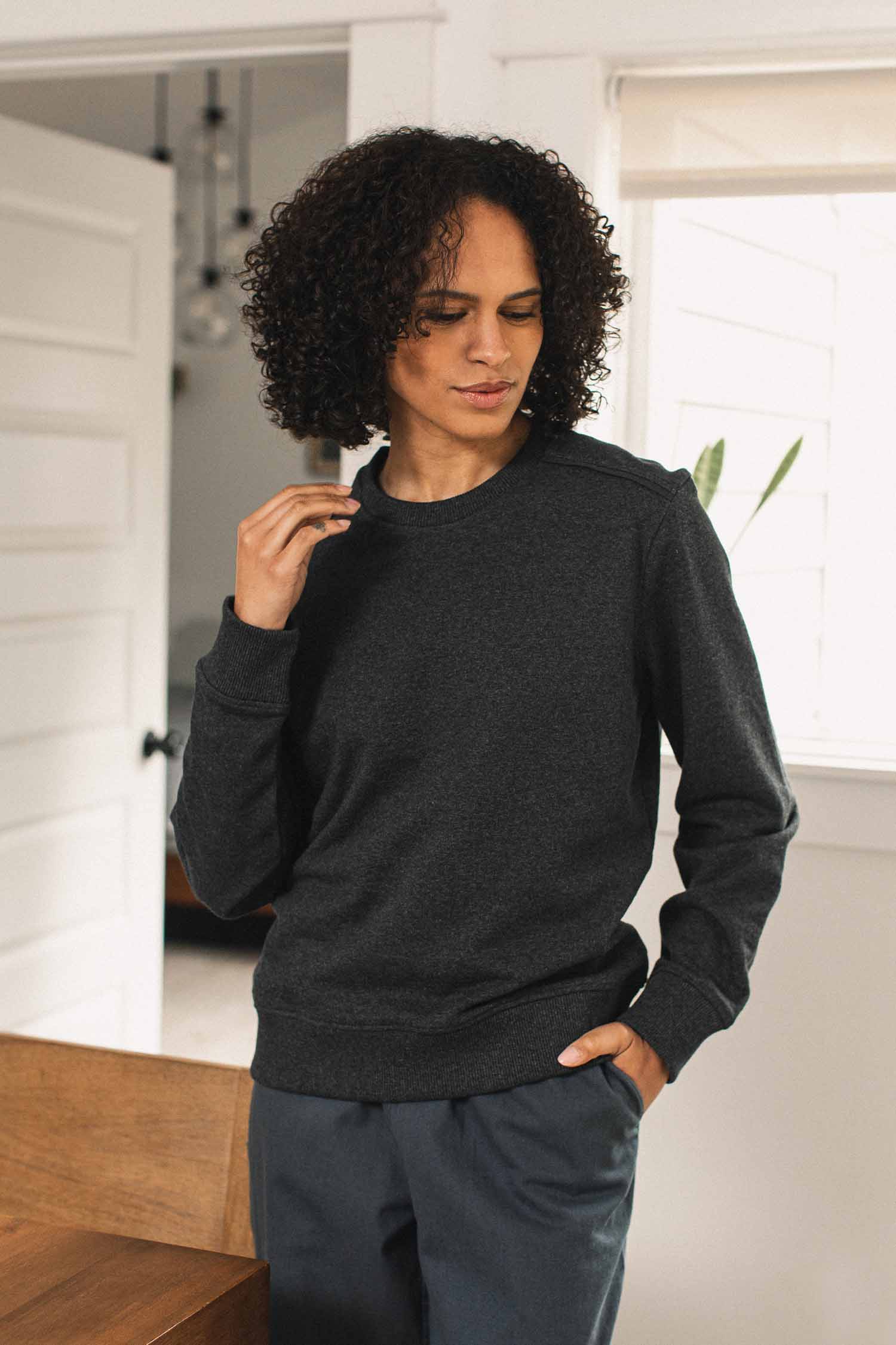 A person with curly hair stands indoors in bright natural light, thoughtfully looking down with one hand in their pocket, wearing the eco-friendly Bridge & Burn Bailey Crew Sweatshirt in Graphite and matching pants.