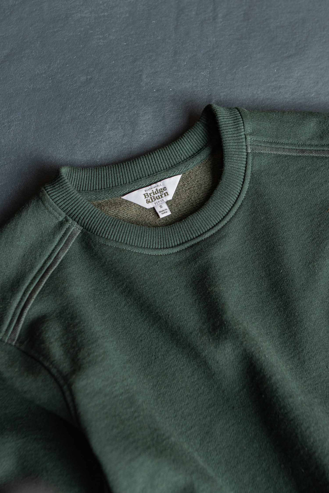 A close-up of the Bridge & Burn Bailey Crew Sweatshirt in Juniper, laid flat on a gray surface, highlighting the collar, shoulder seam, and the interior neck tag.