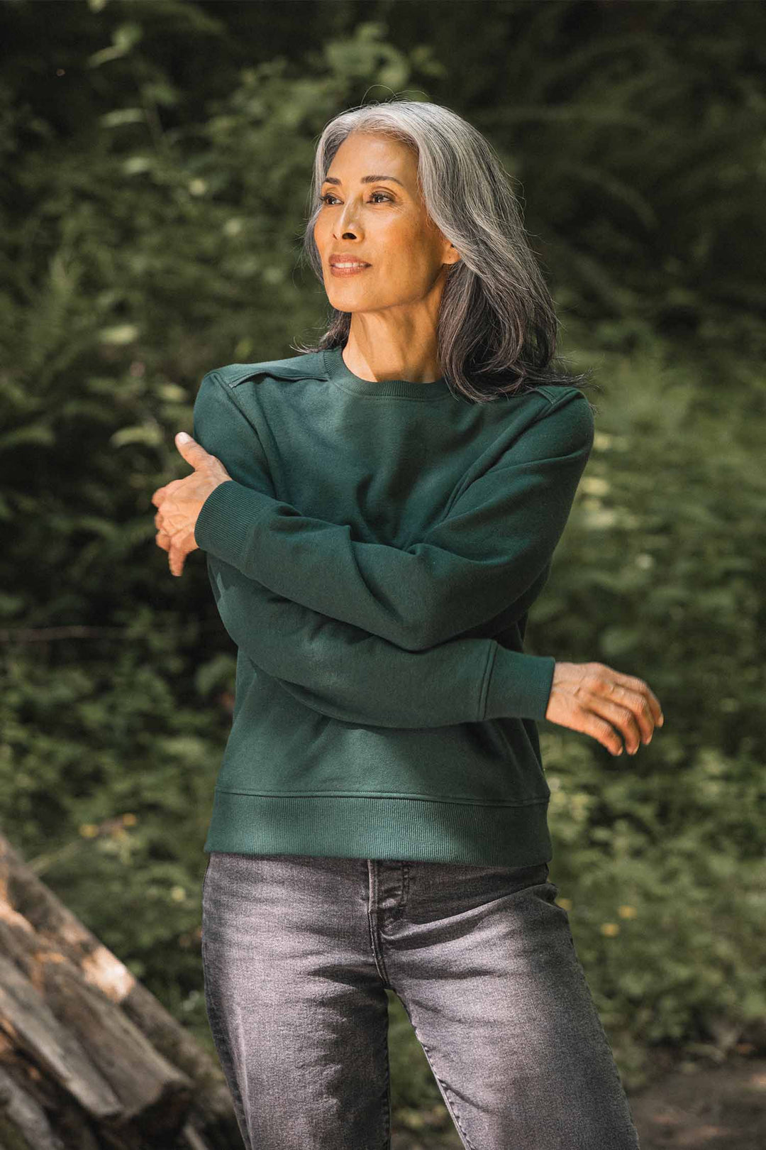 A woman with long gray hair stands outdoors with arms crossed, gazing to the side. She wears the Bridge & Burn Bailey Crew Sweatshirt in Juniper and gray jeans, surrounded by lush greenery bathed in sunlight.