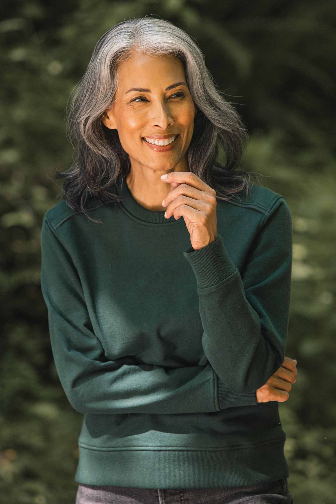 A smiling older woman with long gray and black hair stands outdoors in a forest, wearing the Bridge & Burn Bailey Crew Sweatshirt in Juniper, and holds one hand near her chin. She appears relaxed and content.