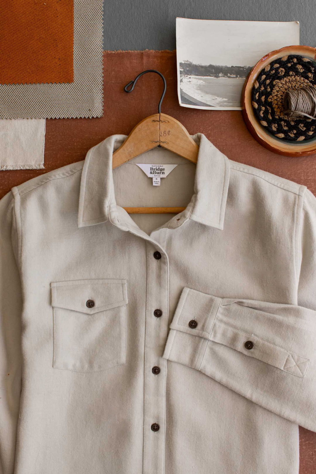 A cream-colored, relaxed fit button-up—Bridge & Burn’s Bird Shirt in Fawn Flannel—hangs on a wooden hanger atop brown fabric, surrounded by fabric swatches, a vintage photo, and a bowl filled with twine and decorative tape. Crafted from organic cotton, this shirt brings effortless style to any wardrobe.