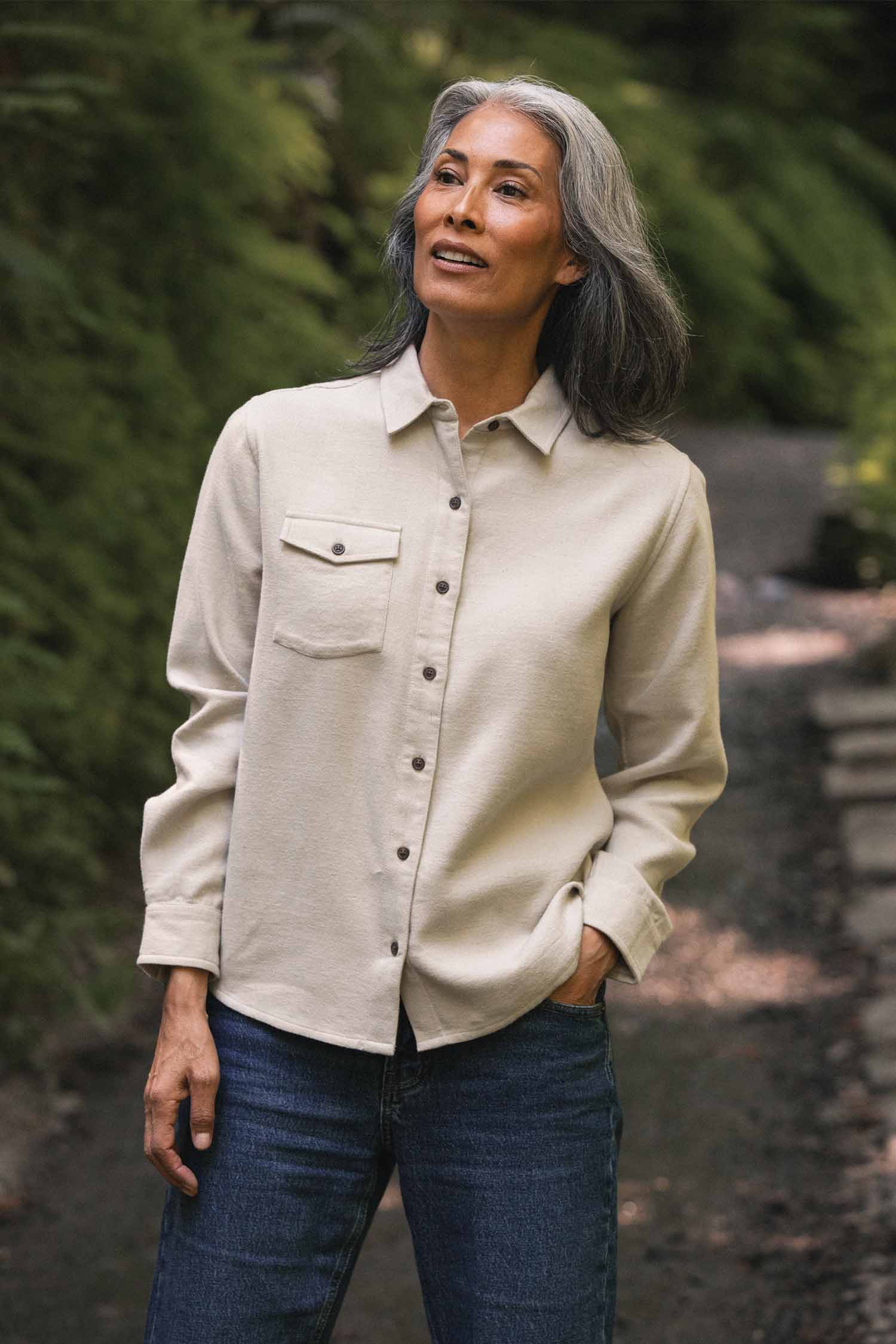 A woman with long gray hair, wearing a relaxed fit Bridge & Burn Bird Shirt in Fawn Flannel made from organic cotton along with blue jeans, stands outdoors on a forest path surrounded by greenery, looking slightly upward and smiling.