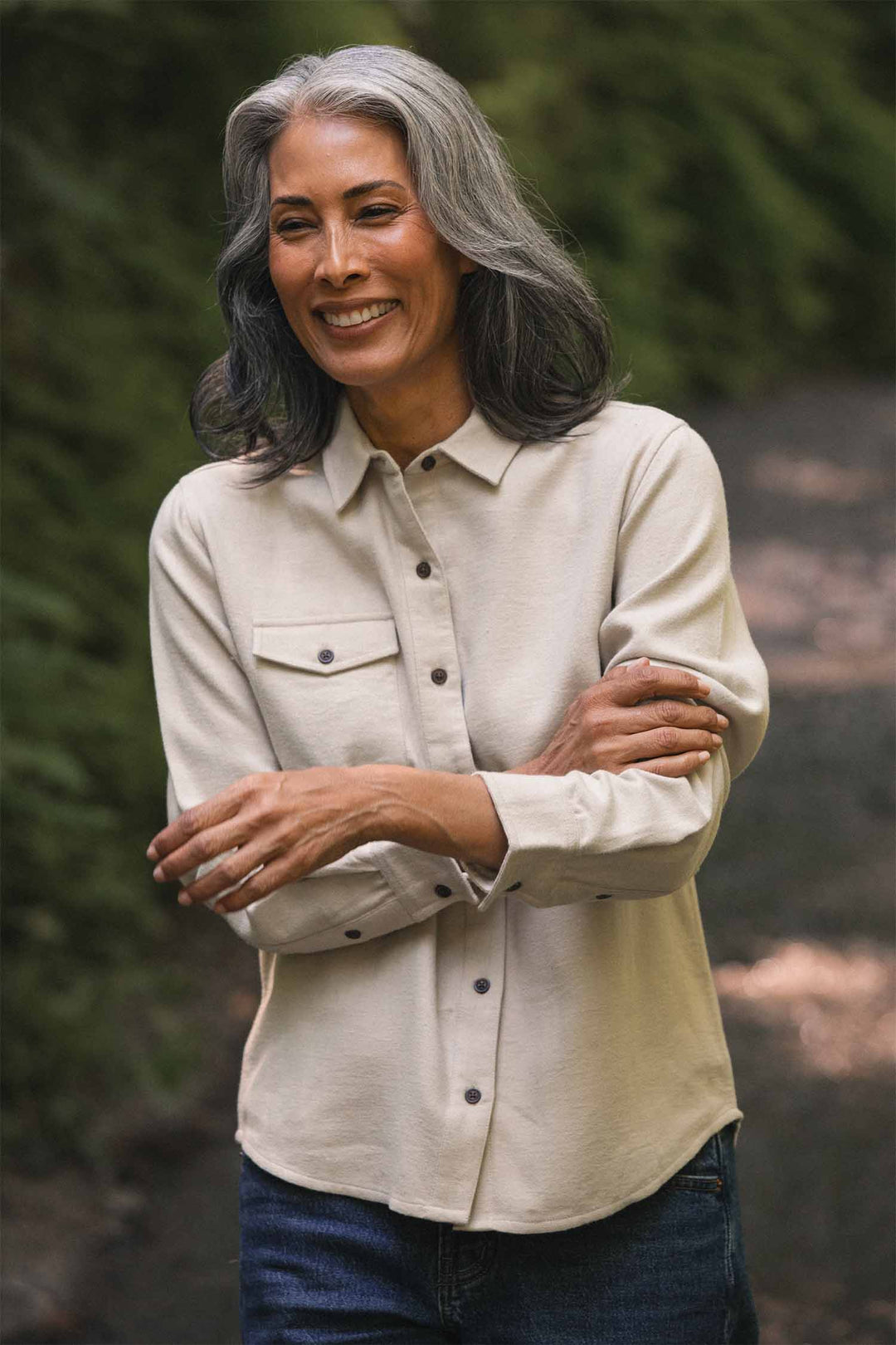 A smiling woman with long, wavy gray hair stands outdoors on a path, wearing the Bridge & Burn Bird Shirt in Fawn Flannel—a relaxed fit made from organic cotton—paired with jeans and her arms crossed. Lush green foliage fills the background.