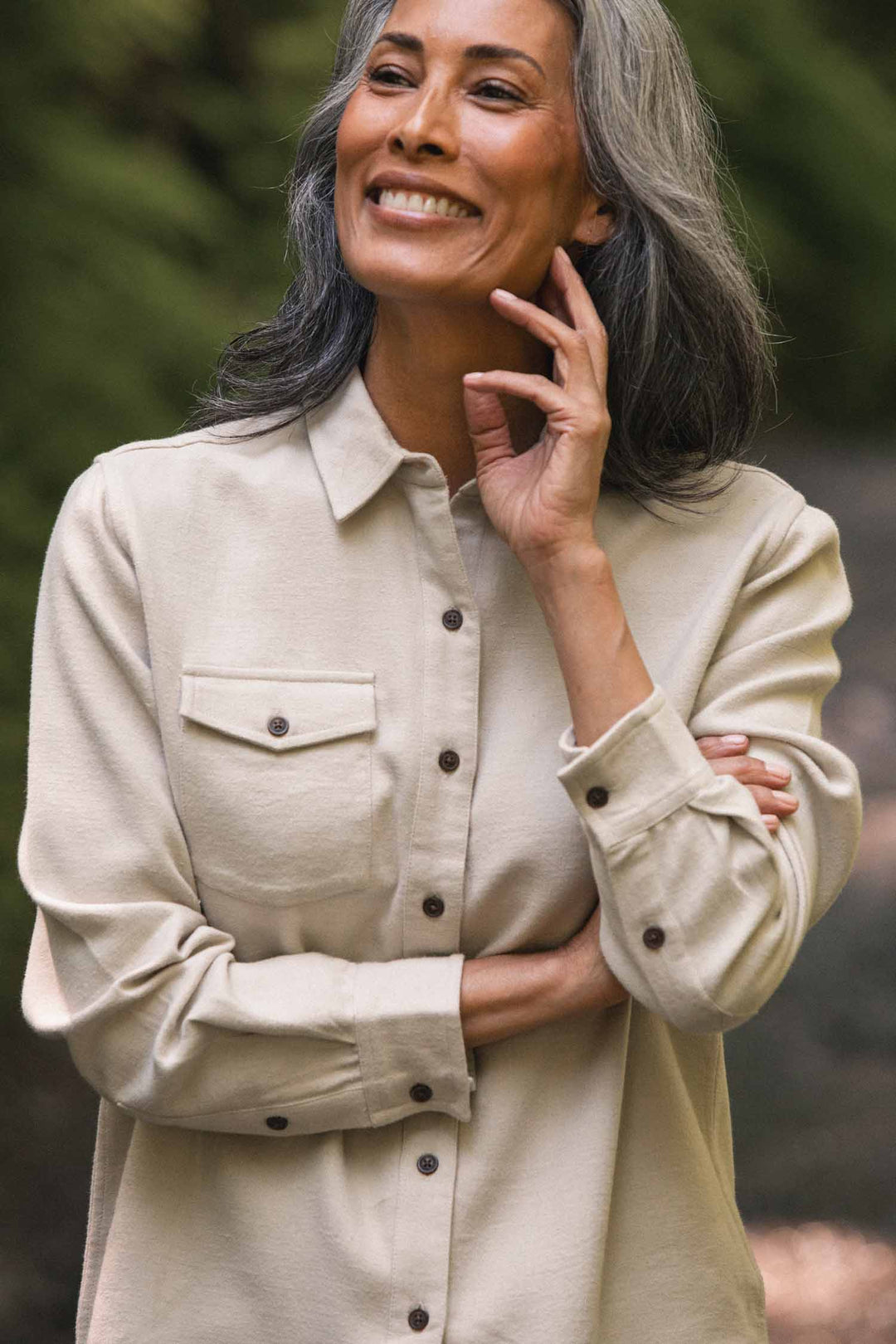 A woman with long gray hair smiles outdoors, wearing the Bridge & Burn Bird Shirt in Fawn Flannel—a relaxed fit, organic cotton top featuring chest pockets. She holds one hand near her face and crosses her other arm, standing against a blurred green background.