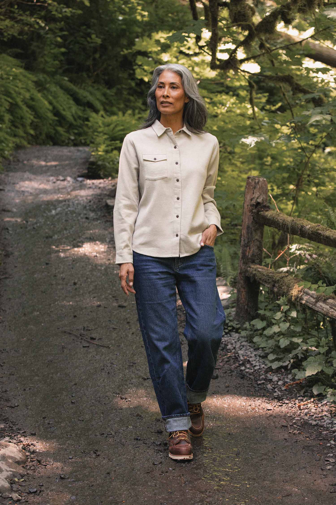 A person with gray hair walks on a forest path, wearing the Bridge & Burn Bird Shirt in Fawn Flannel—a relaxed fit, cream-colored shirt made from organic cotton—paired with blue jeans and brown boots. Lush green foliage and a wooden fence line the trail.