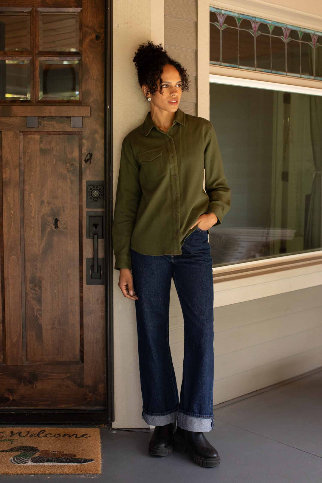 A woman wearing the Bridge & Burn Bird Shirt in Pine Flannel—a relaxed-fit, organic cotton green button-up—paired with rolled-up jeans stands on a porch near a brown wooden door, looking to the side. A “Welcome” mat lies at her feet.