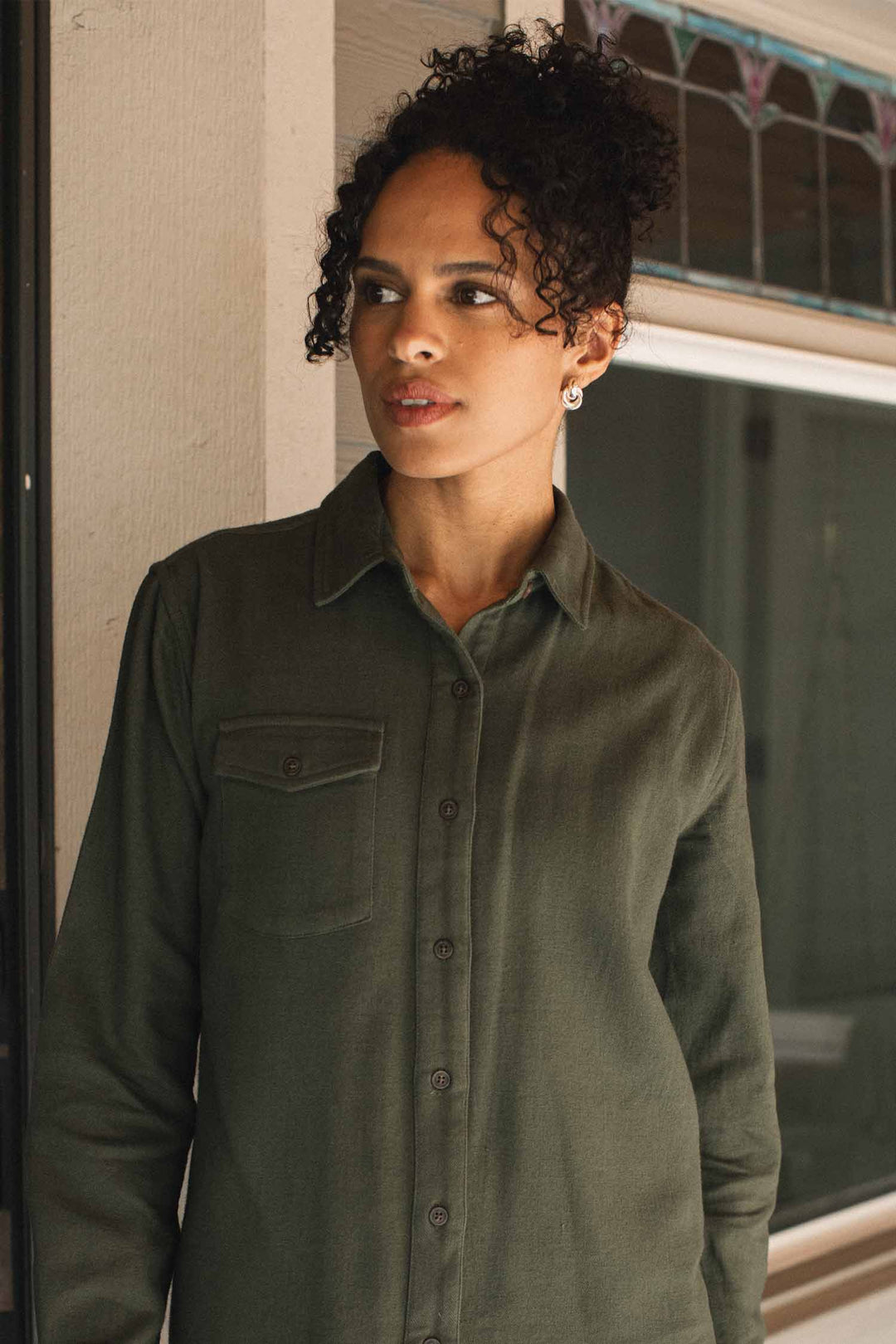 A person with curly hair, wearing hoop earrings and the Bridge & Burn Bird Shirt in Pine Flannel—a relaxed-fit, organic cotton green button-up—stands indoors near a window with stained glass details.