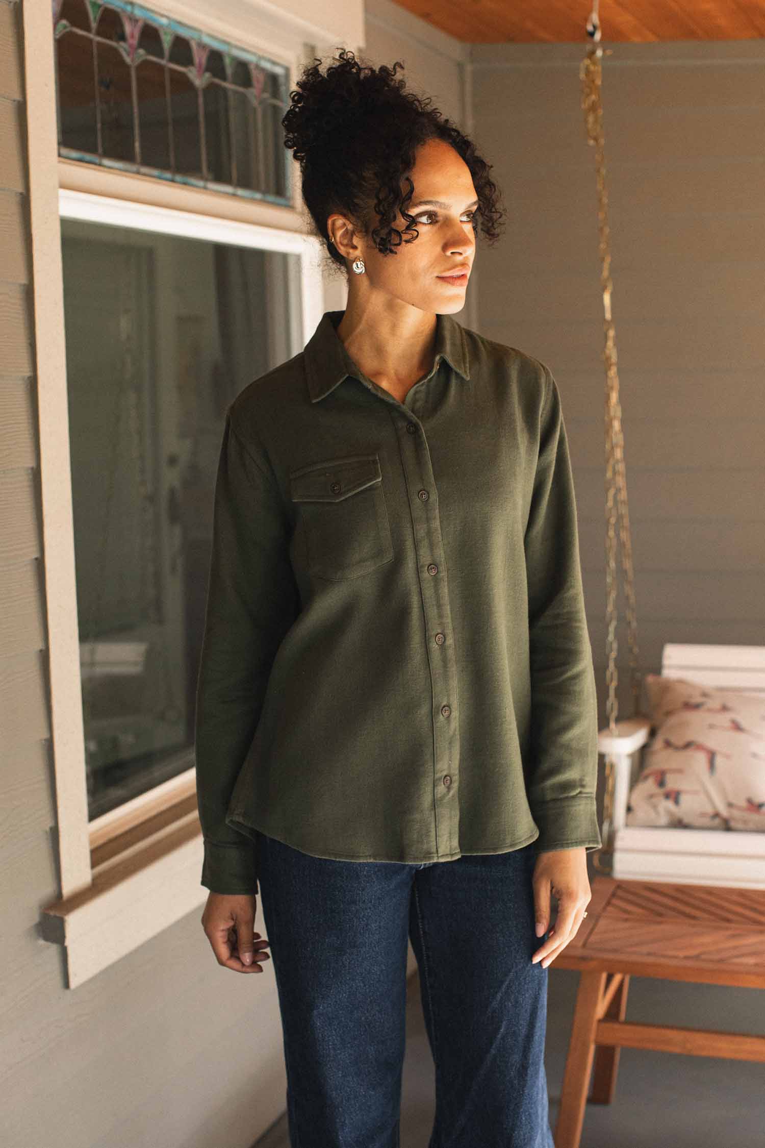 Wearing the Bridge & Burn Bird Shirt in Pine Flannel—a relaxed fit, olive green button-up made from organic cotton—a woman with curly hair stands on a porch beside a window and a bench with pillows, paired with dark jeans.