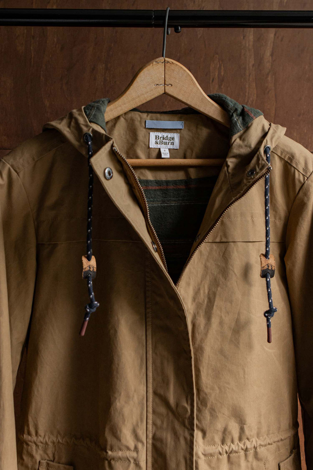 Bridge & Burn Briar Jacket / Dark Khaki Waxed Canvas hooded parka with a front zipper, snap closure, and dark patterned drawstrings, hanging on a wooden hanger.