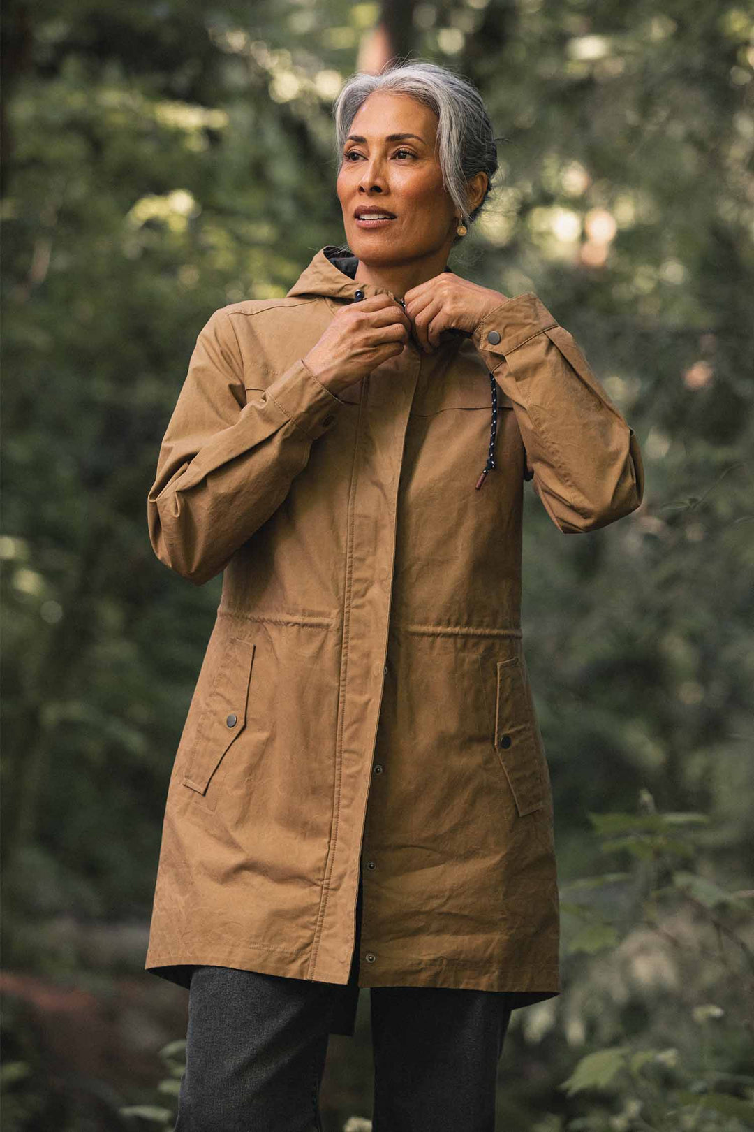 A person wearing a Bridge & Burn Briar Jacket / Dark Khaki Waxed Canvas, a hooded, khaki-colored utility parka with a front zipper and snap buttons.