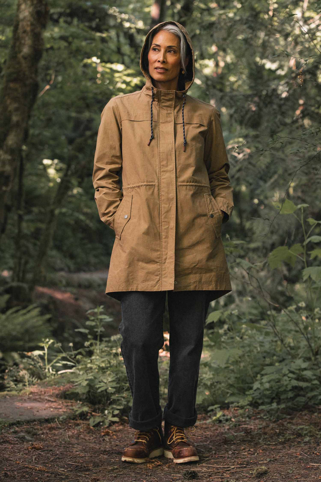A person wearing a hooded, khaki-colored Bridge & Burn Briar Jacket / Dark Khaki Waxed Canvas parka in a wooded area.