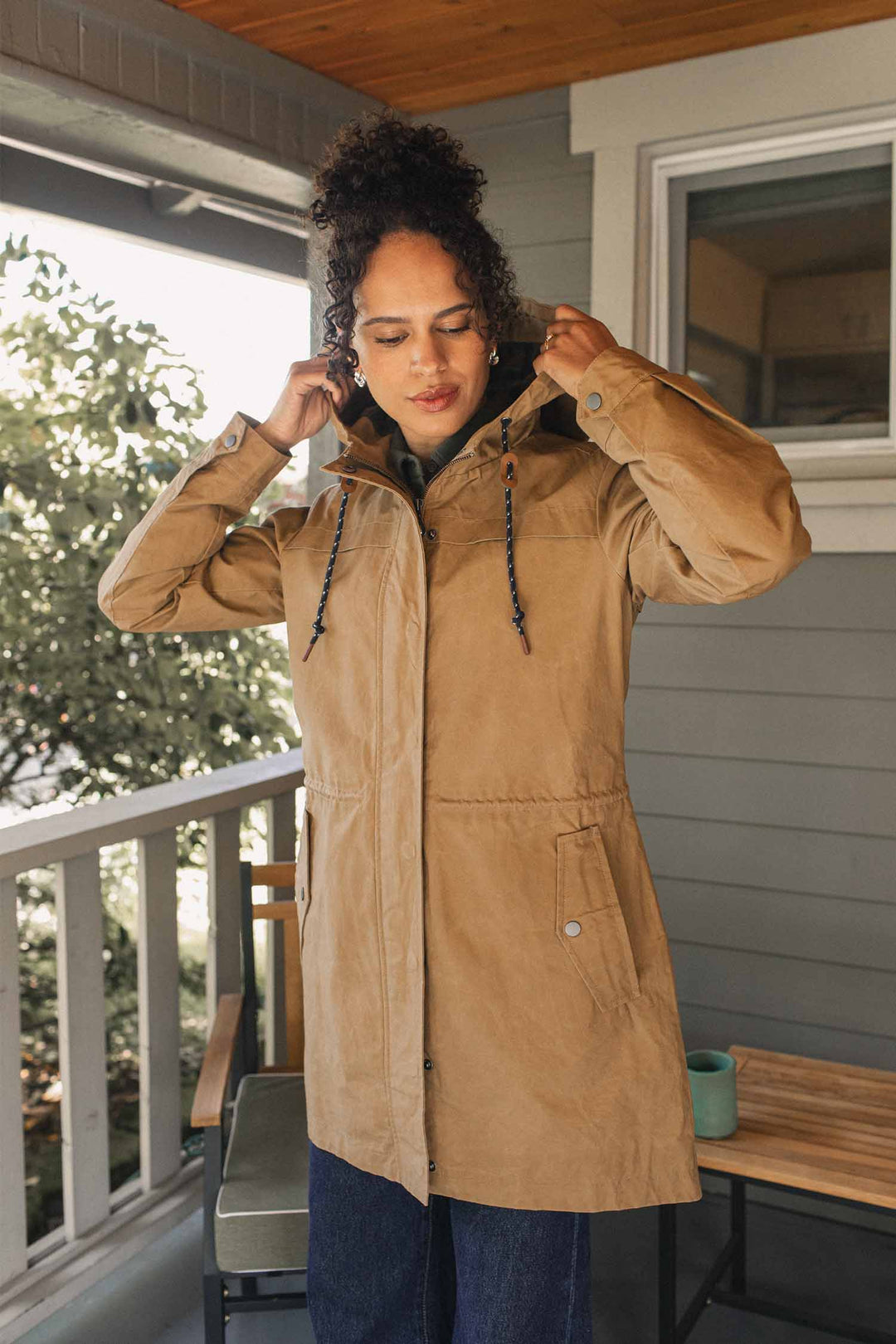 A woman wearing a tan Bridge & Burn Briar Jacket / Dark Khaki Waxed Canvas, a rain-ready hooded utility parka with a front zipper and snap pockets.