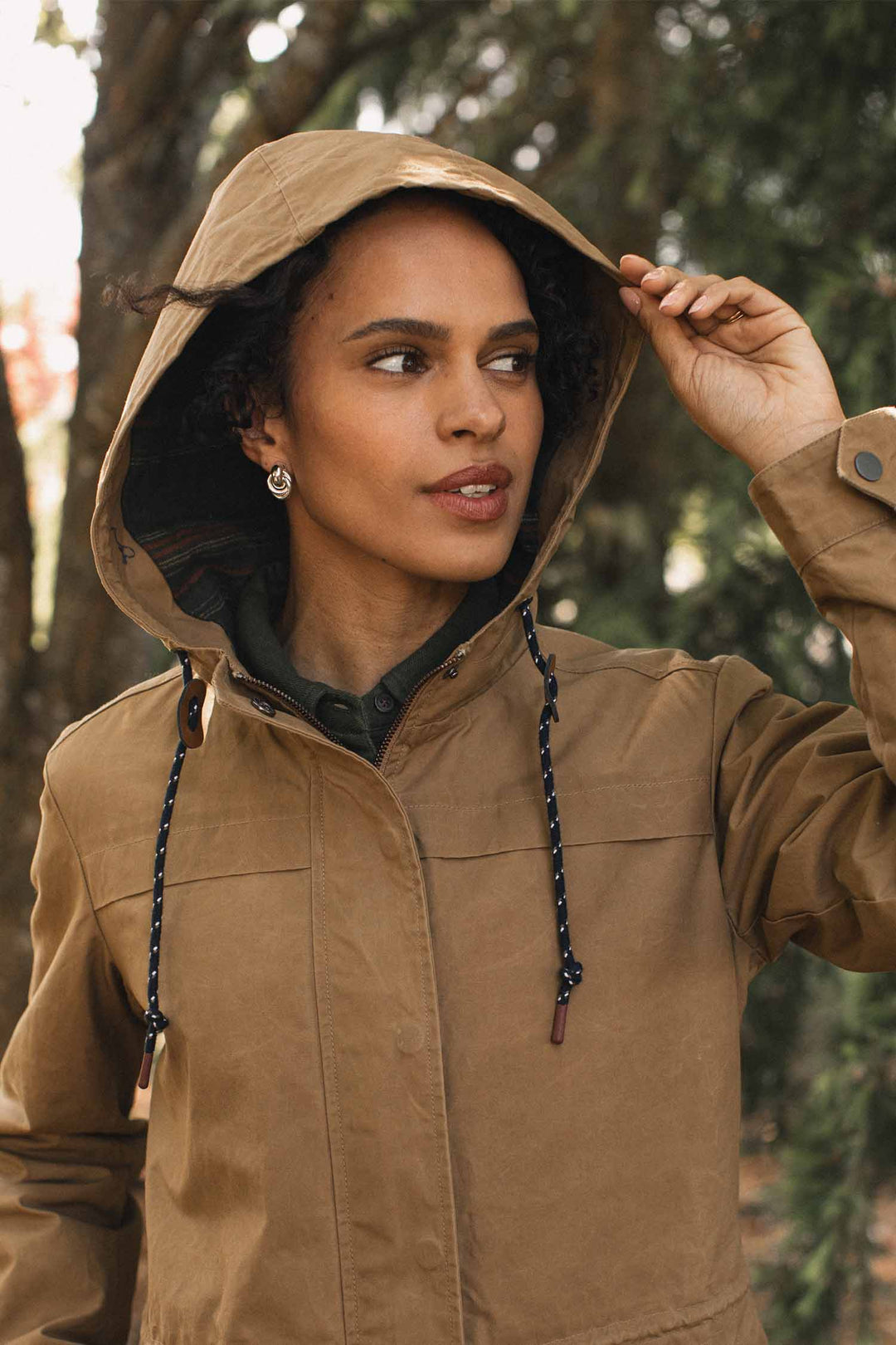 A person wearing a Briar Jacket / Dark Khaki Waxed Canvas, a hooded parka in khaki British Millerain waxed cotton featuring a patterned lining and navy-and-white drawstring cords.