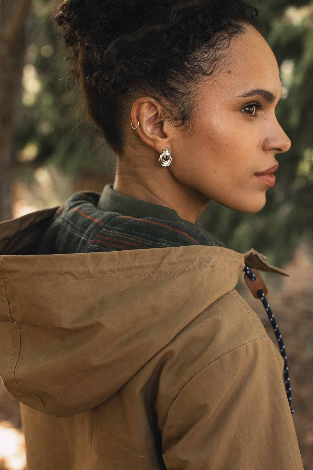 A person seen from the side, wearing a khaki Bridge & Burn Briar Jacket / Dark Khaki Waxed Canvas hooded parka over a green plaid shirt.
